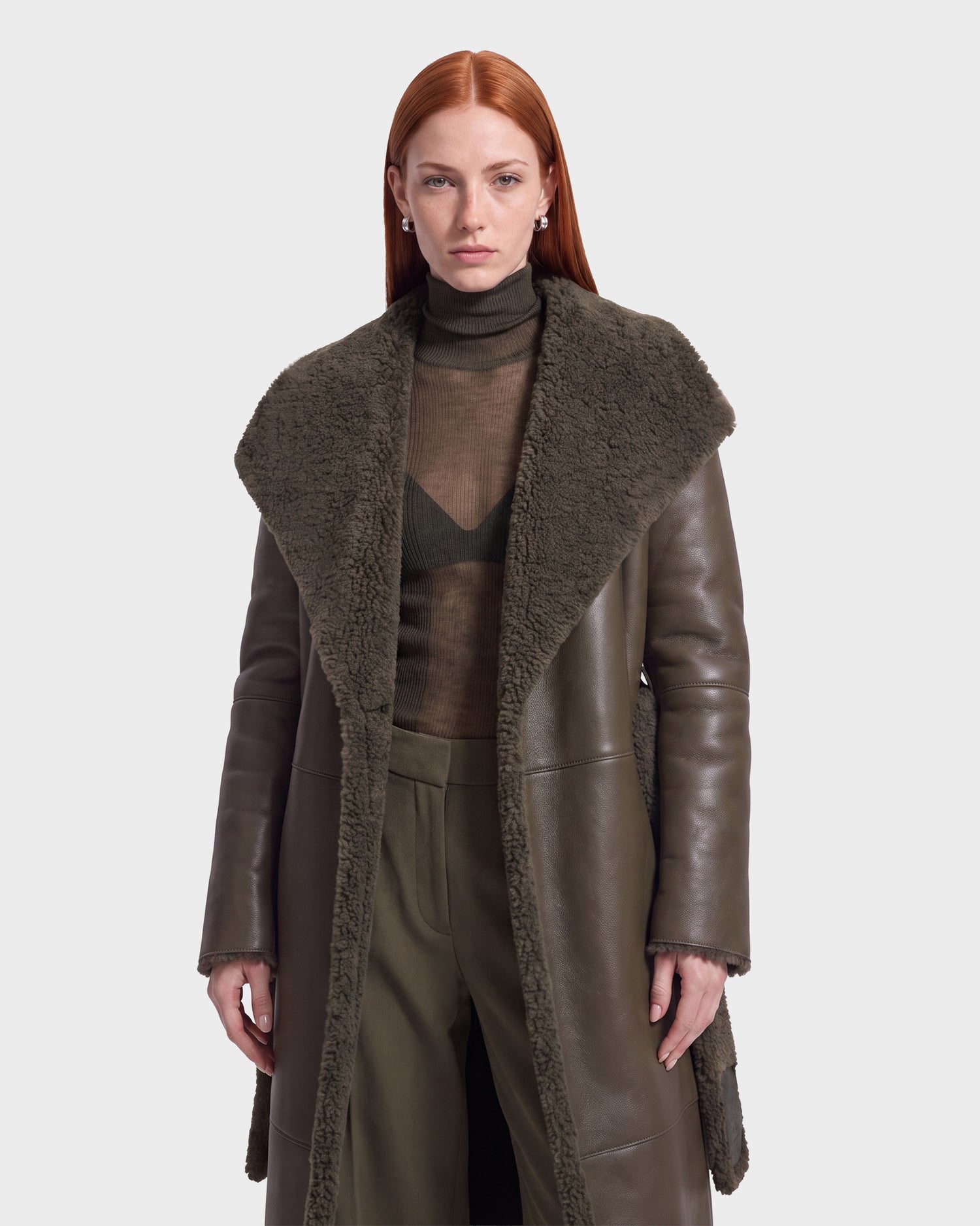 A woman with straight red hair wears the FRANCIE - 8126873 Olive shearling coat by Rudsak over a sheer turtleneck and matching pants, standing against a plain light background. Available for pre-order now, this look ships September 15.