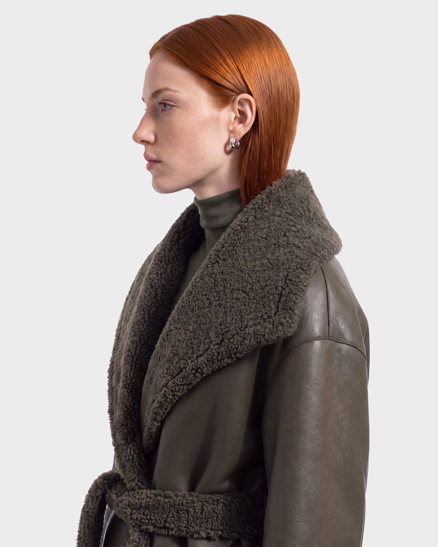 A person with straight red hair, wearing the Rudsak FRANCIE - 8126873 Olive dark shearling coat and silver earrings, is shown in profile against a plain light background. This striking look is now available for pre-order and ships September 15.