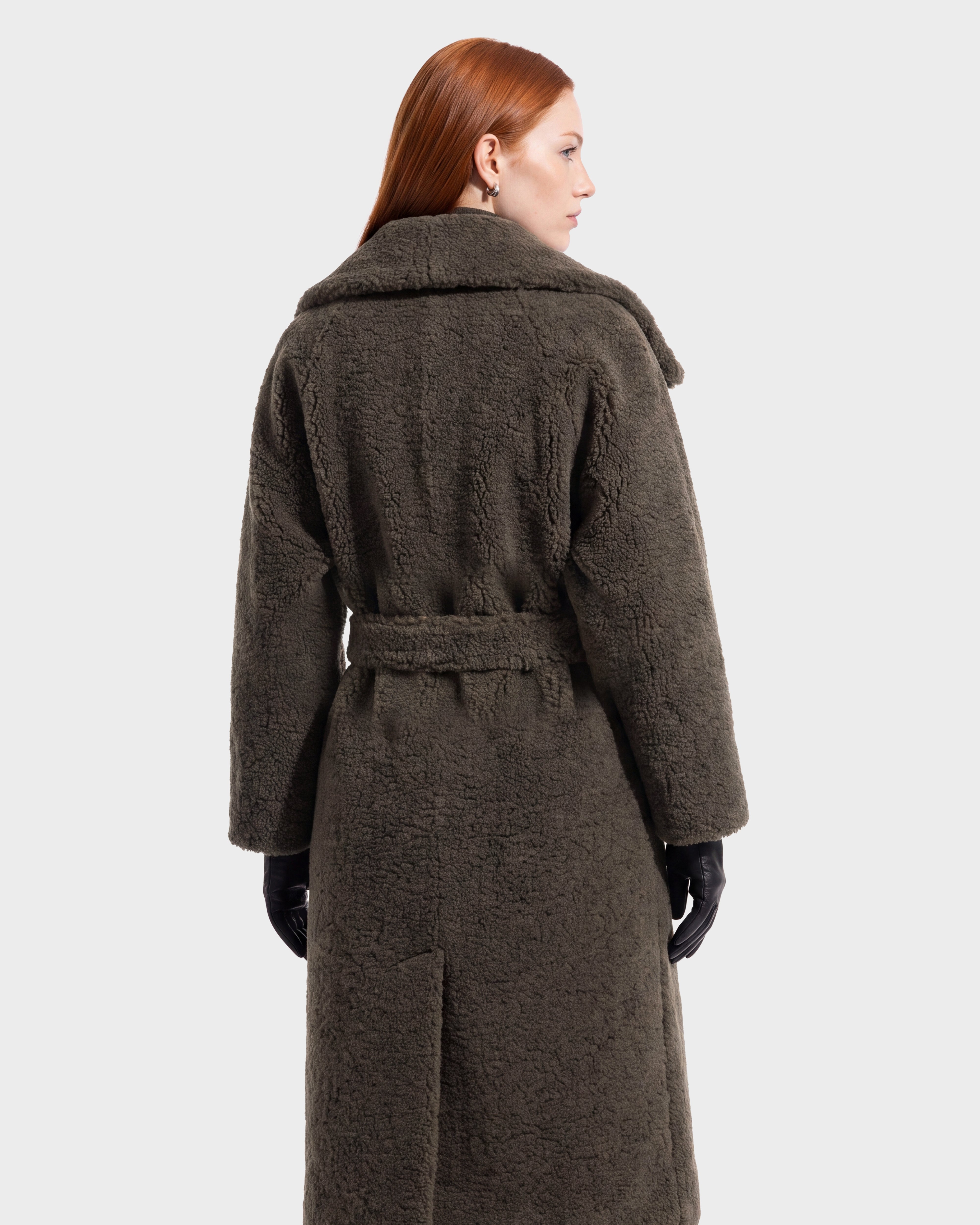 A person with long red hair models the FRANCIE - 8126873 Olive coat by Rudsak—a long, dark textured piece featuring a waist belt—seen from the back against a plain light background. This stunning coat is available for pre-order and is expected to ship by September 15th.