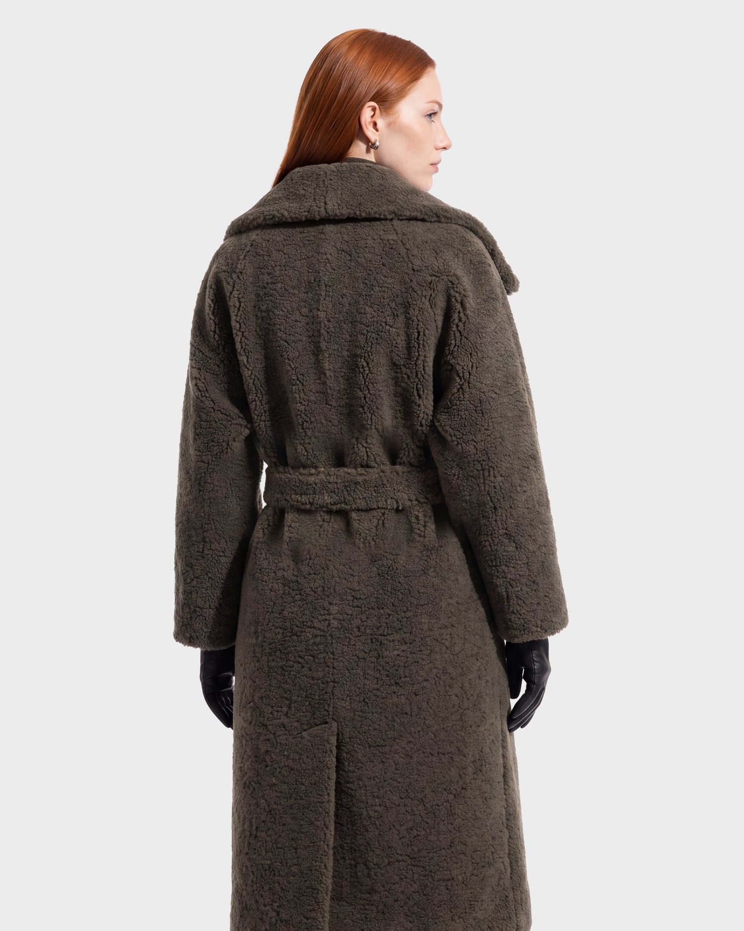 A person with long red hair models the FRANCIE - 8126873 Olive coat by Rudsak—a long, dark textured piece featuring a waist belt—seen from the back against a plain light background. This stunning coat is available for pre-order and is expected to ship by September 15th.