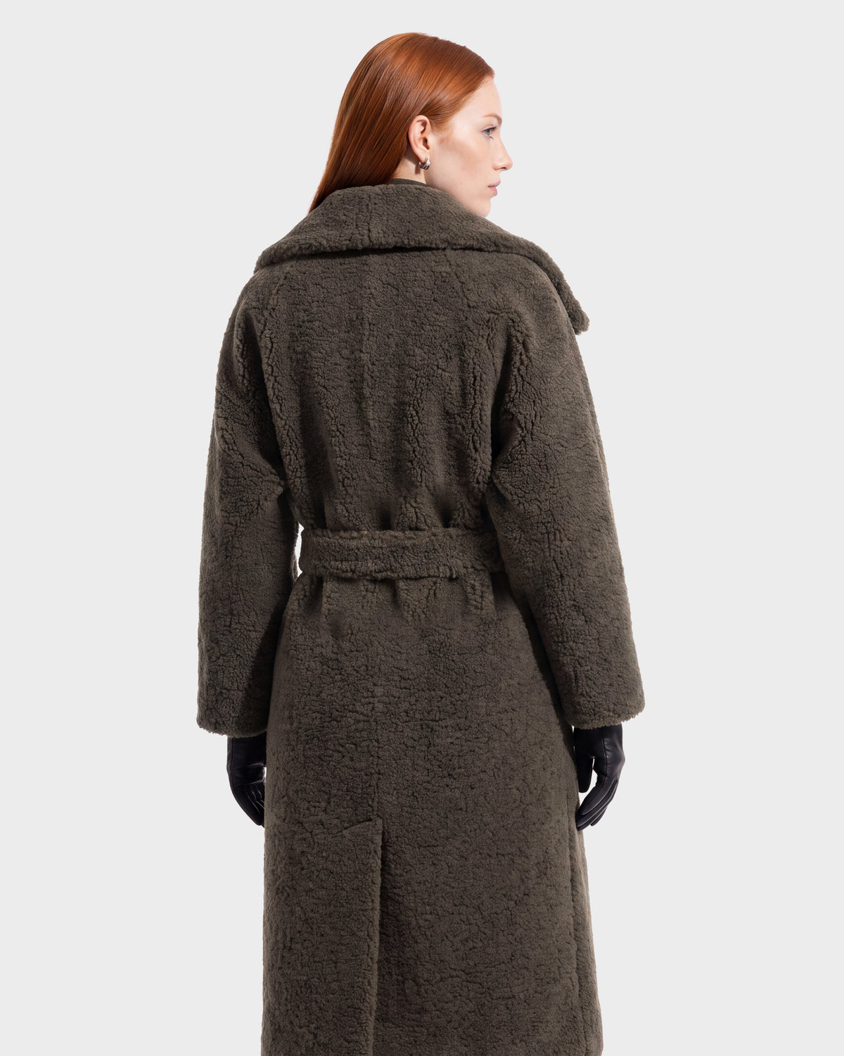 A person with long red hair models the FRANCIE - 8126873 Olive coat by Rudsak—a long, dark textured piece featuring a waist belt—seen from the back against a plain light background. This stunning coat is available for pre-order and is expected to ship by September 15th.