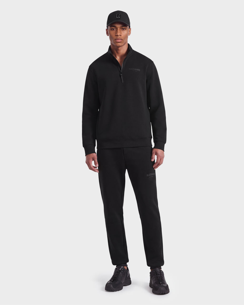 A man wearing a black cap, limited-edition Harvard THE COOP unisex pullover quarter-zip sweatshirt, black jogger pants, and Rudsak FREMONT HV - H525501 Black sneakers stands facing forward against a plain white background.