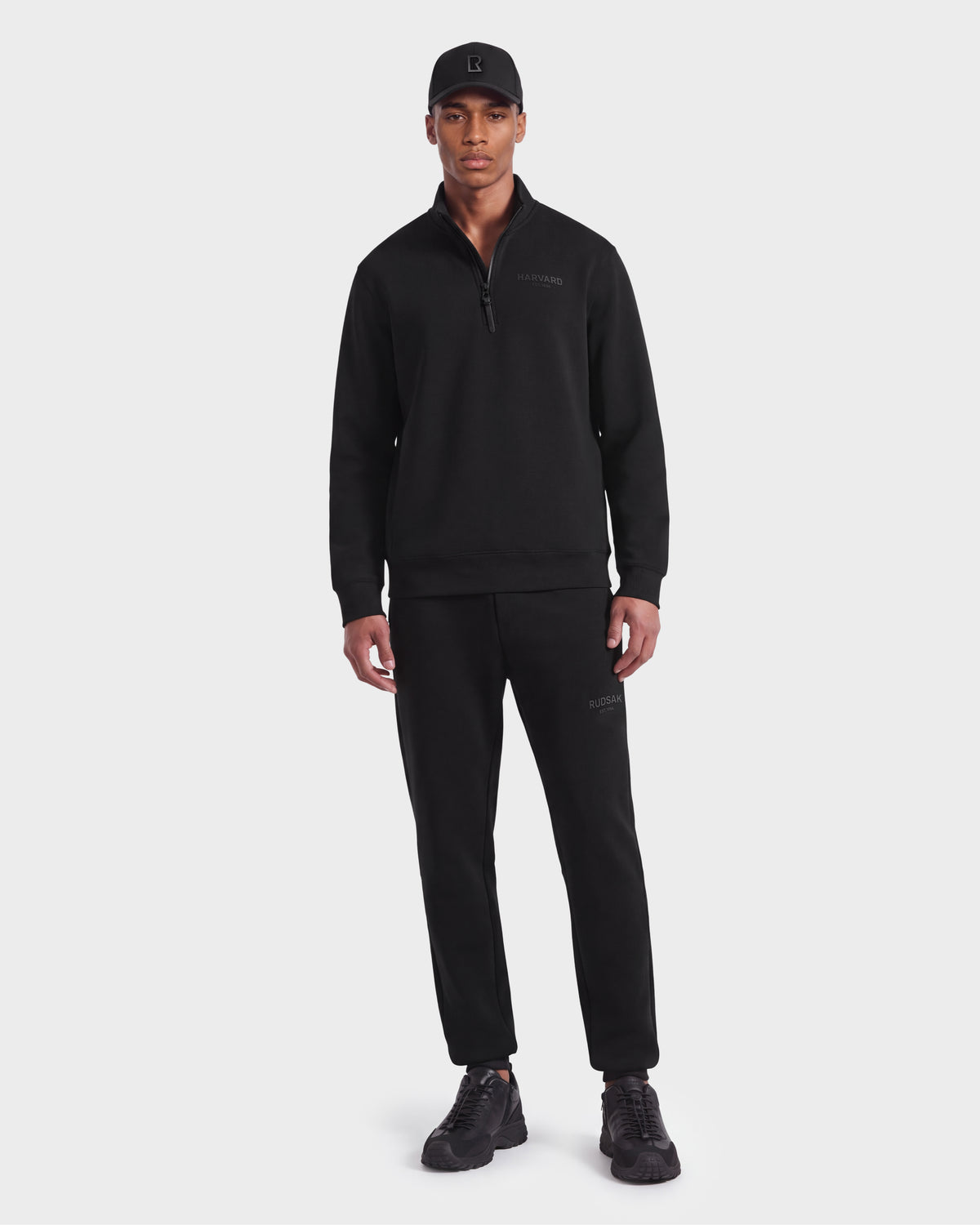 A man wearing a black cap, limited-edition Harvard THE COOP unisex pullover quarter-zip sweatshirt, black jogger pants, and Rudsak FREMONT HV - H525501 Black sneakers stands facing forward against a plain white background.