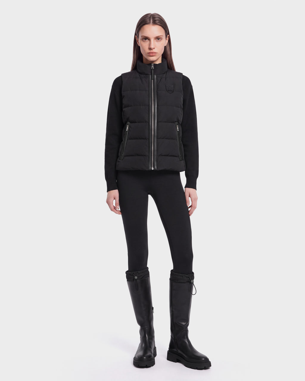 A woman with straight brown hair and a neutral expression stands against a white background, wearing the Rudsak GABRIELA HV - H125507 Black sleek zip-up puffer vest made with RDS-certified down, paired with a black long-sleeve top, black leggings, and knee-high black boots.
