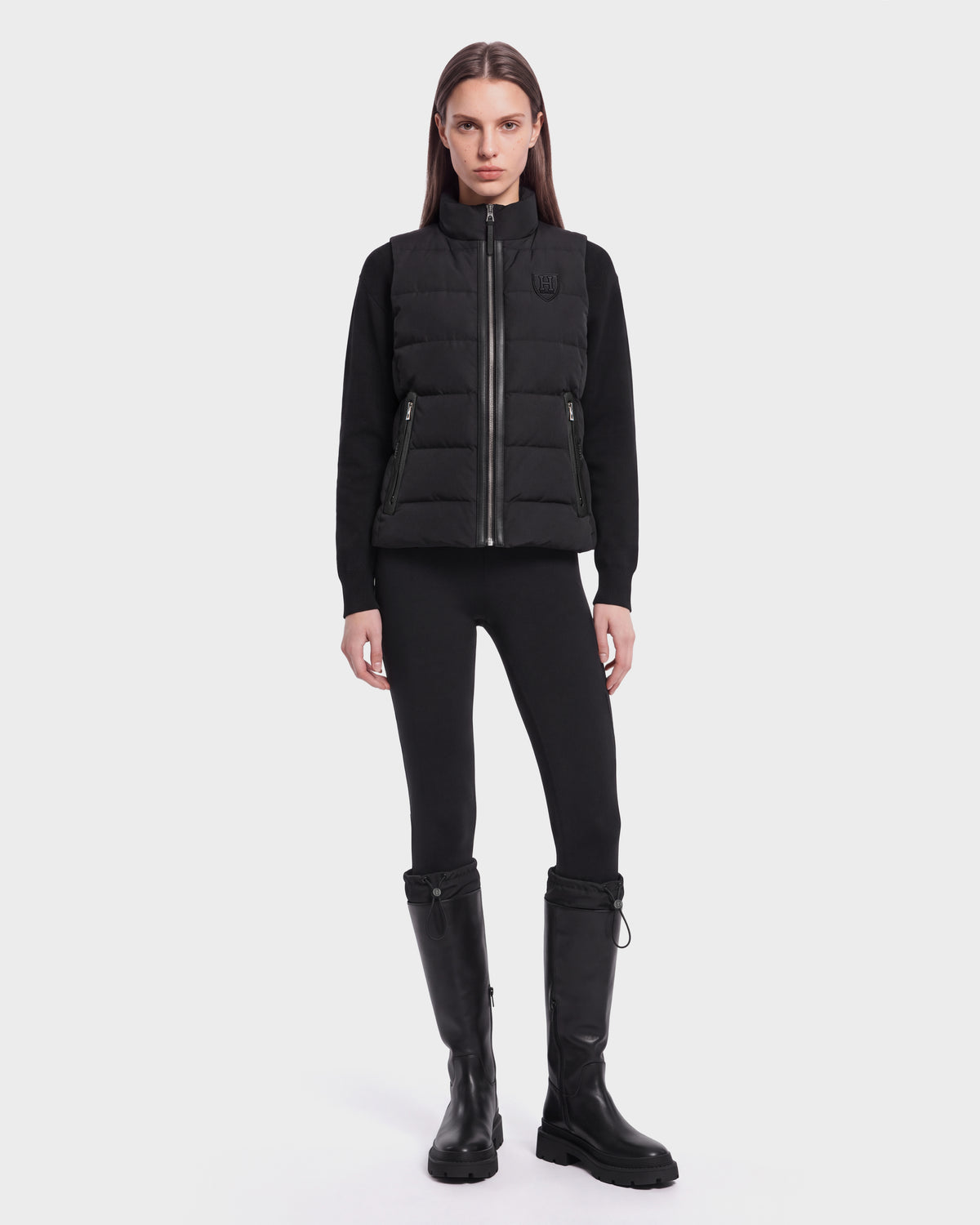 A woman with straight brown hair and a neutral expression stands against a white background, wearing the Rudsak GABRIELA HV - H125507 Black sleek zip-up puffer vest made with RDS-certified down, paired with a black long-sleeve top, black leggings, and knee-high black boots.