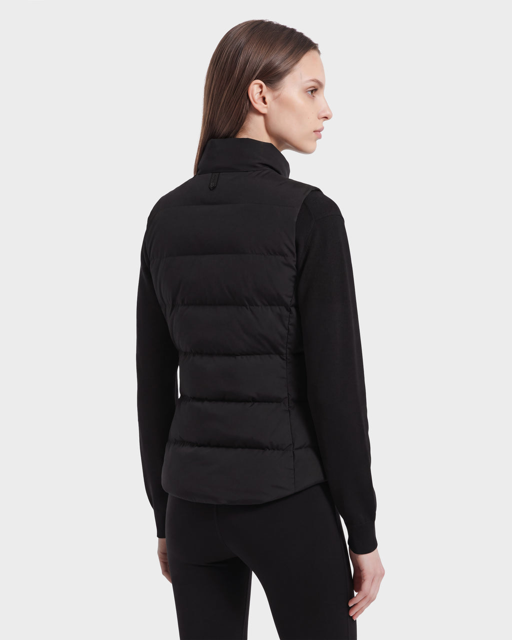A woman with straight brown hair, dressed in the Rudsak GABRIELA HV - H125507 Black quilted puffer vest over a black long-sleeve top, stands facing away and slightly to the side against a plain light background.