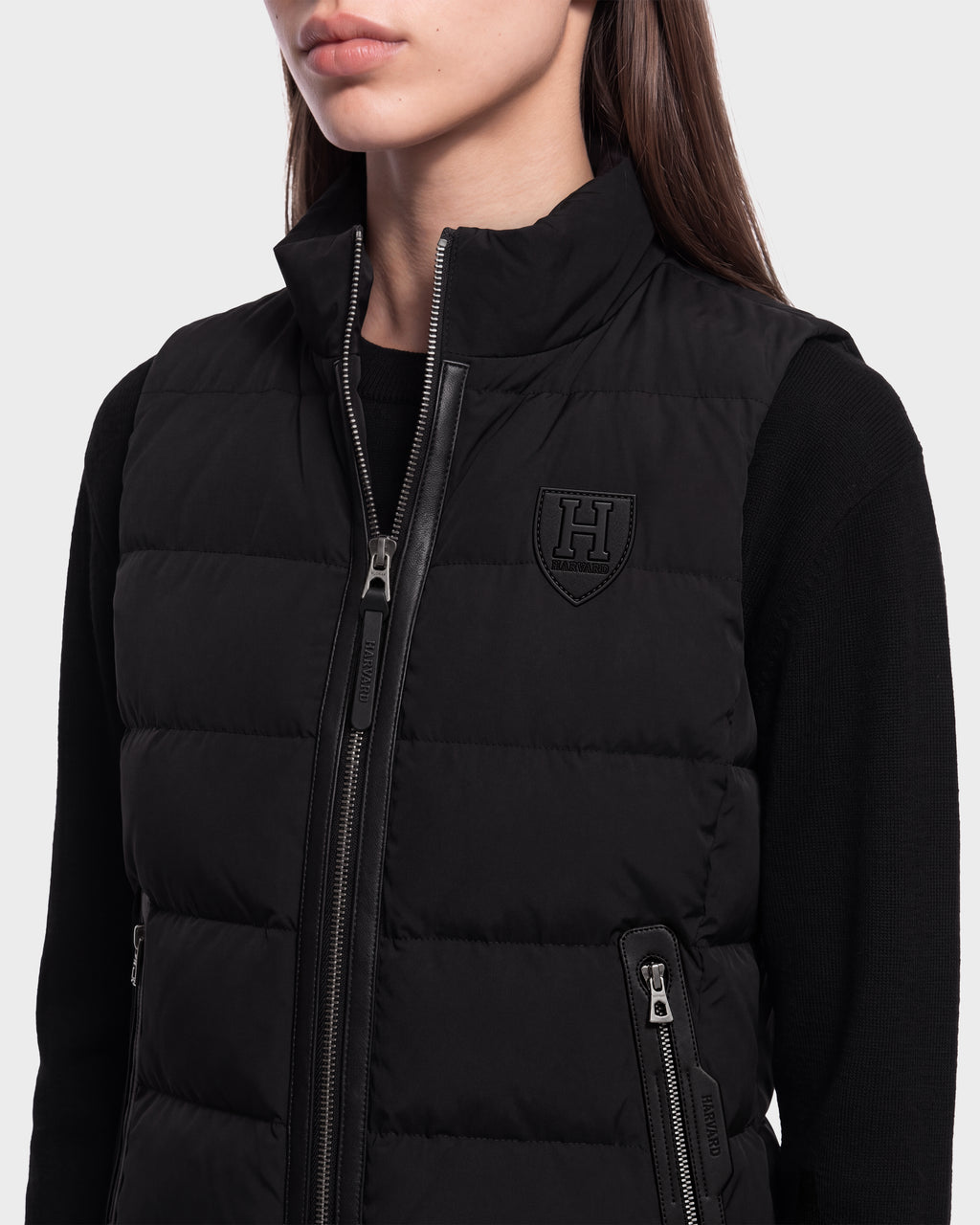 A woman wears the Rudsak GABRIELA HV - H125507 Black vest, a quilted puffer with a zipper and Harvard crest on the chest, layered over a black long-sleeve top and shown from shoulders to mid-torso—a sleek piece of Harvard apparel.