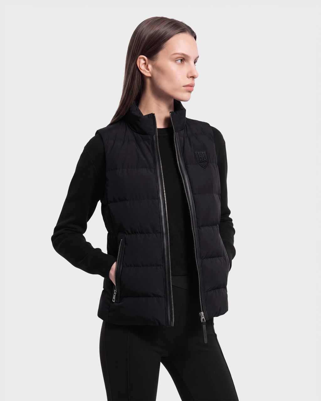 A woman with straight brown hair models the Rudsak GABRIELA HV - H125507 Black RDS-certified down puffer vest, layered over a black long-sleeve shirt and black pants. She stands with one hand in her pocket against a plain light background.