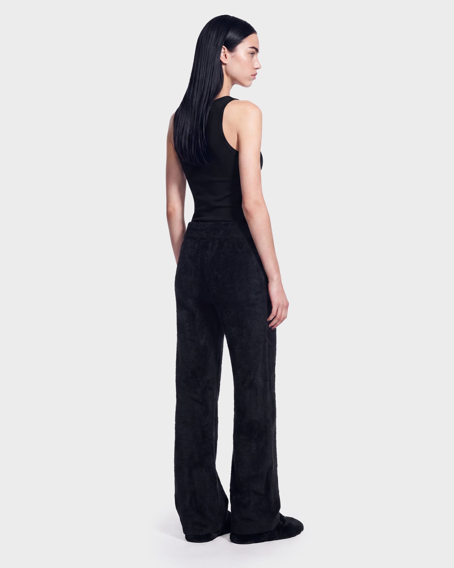 A woman with straight black hair is wearing the Rudsak GENESIS - 8526506 Black sleeveless top and matching black wide-leg pants, standing and facing slightly away against a plain light background.