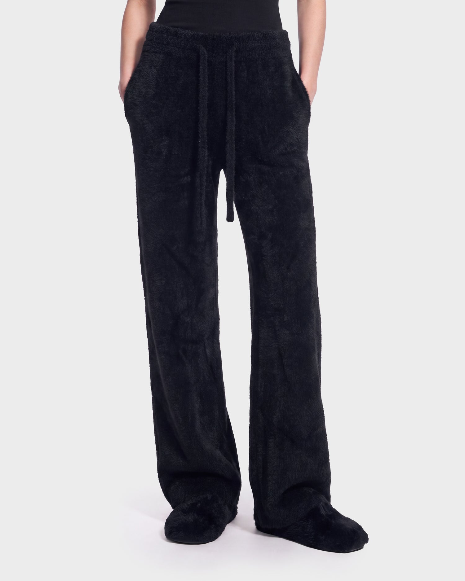 A person is shown from the waist down wearing Rudsak’s GENESIS - 8526506 Black lounge pants—black, fuzzy, and loose-fitting with a drawstring waist and matching attached slippers—standing against a plain light background.