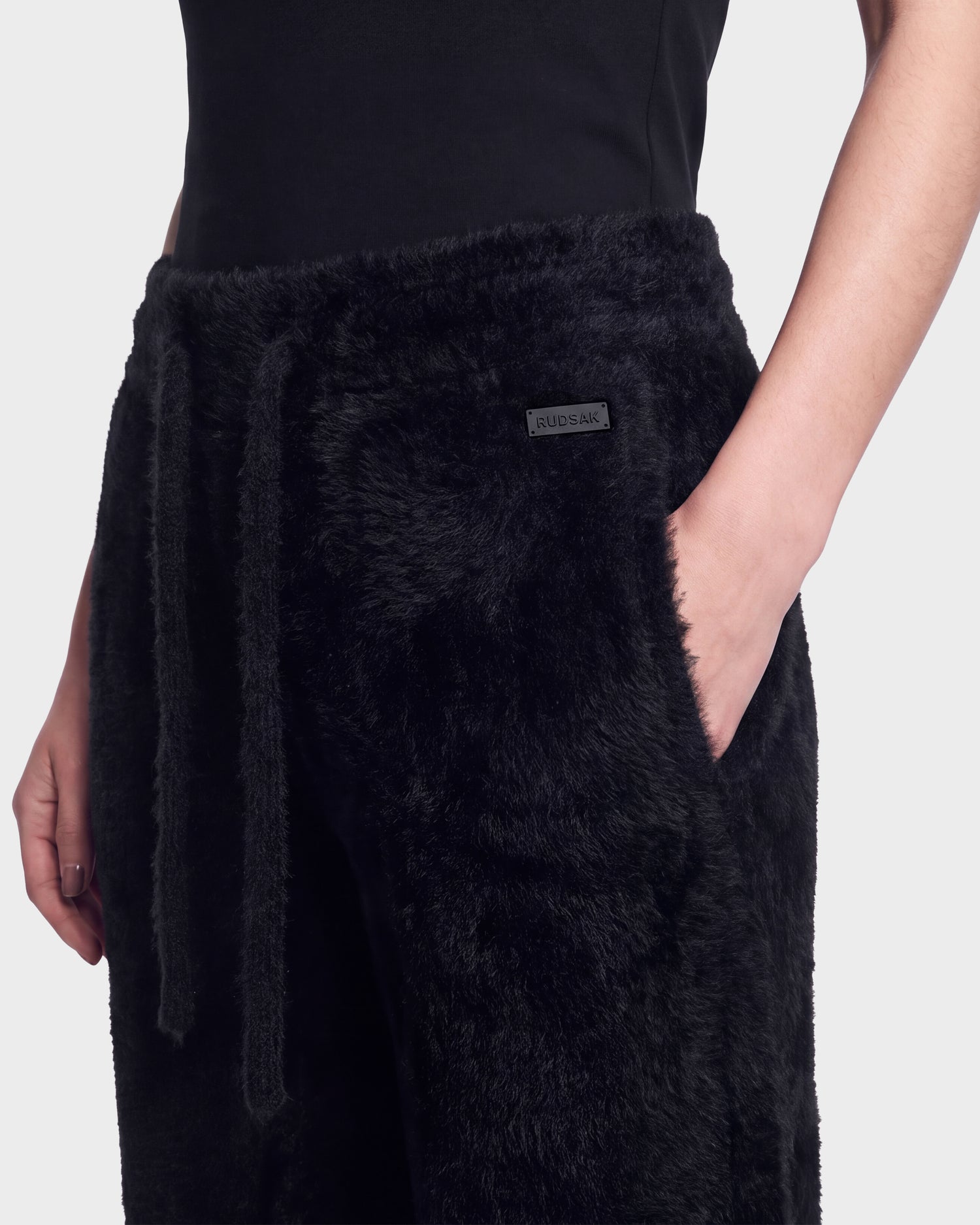 A person wearing the Rudsak GENESIS - 8526506 Black fuzzy pants with a drawstring and a small rectangular label on the waistband, paired with a black top. One hand is in the pocket.