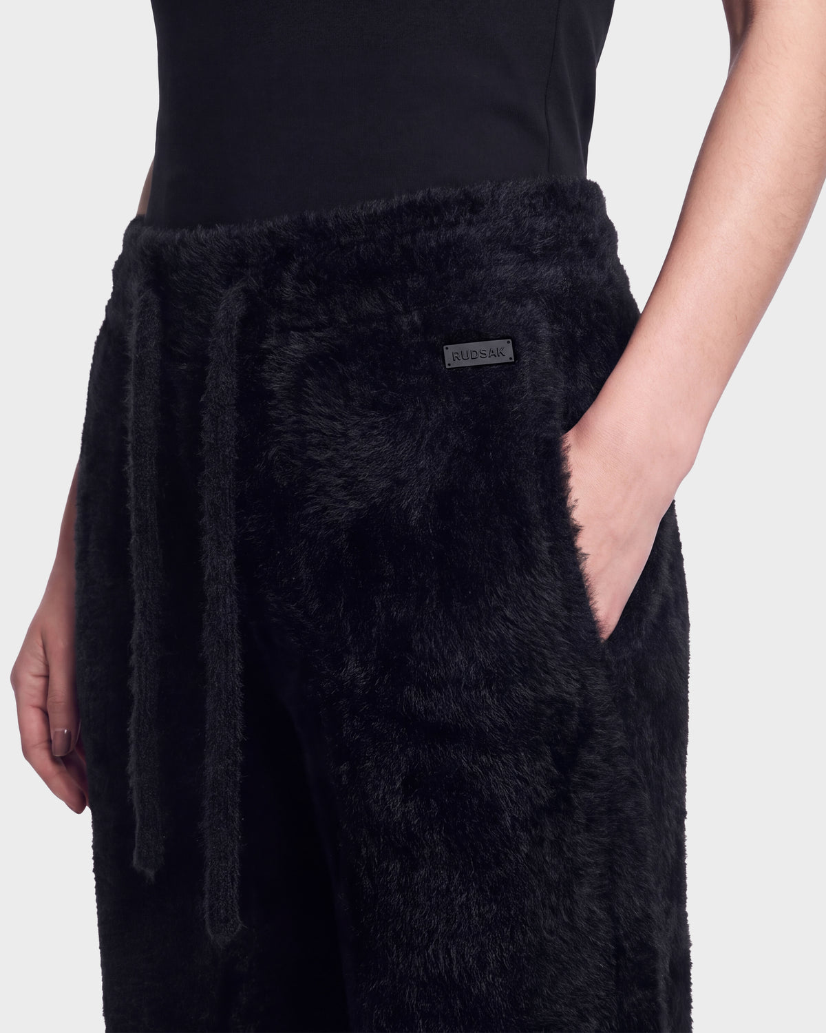 A person wearing the Rudsak GENESIS - 8526506 Black fuzzy pants with a drawstring and a small rectangular label on the waistband, paired with a black top. One hand is in the pocket.