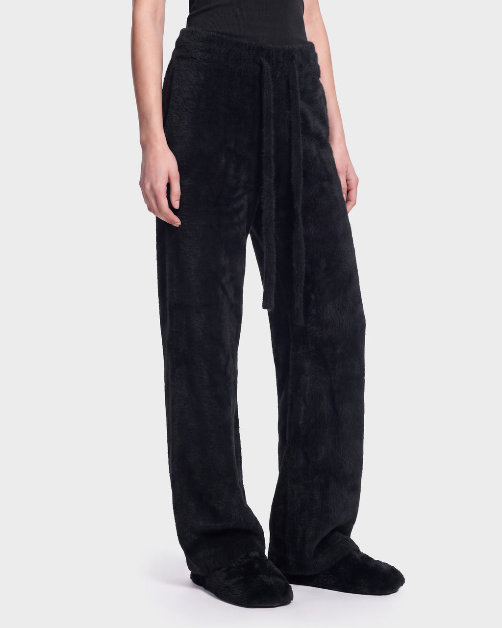 A person is shown from the waist down wearing Rudsak’s GENESIS - 8526506 Black pants—loose-fitting, fuzzy black trousers with a matching tie belt—paired with black slippers, standing against a plain, light background.