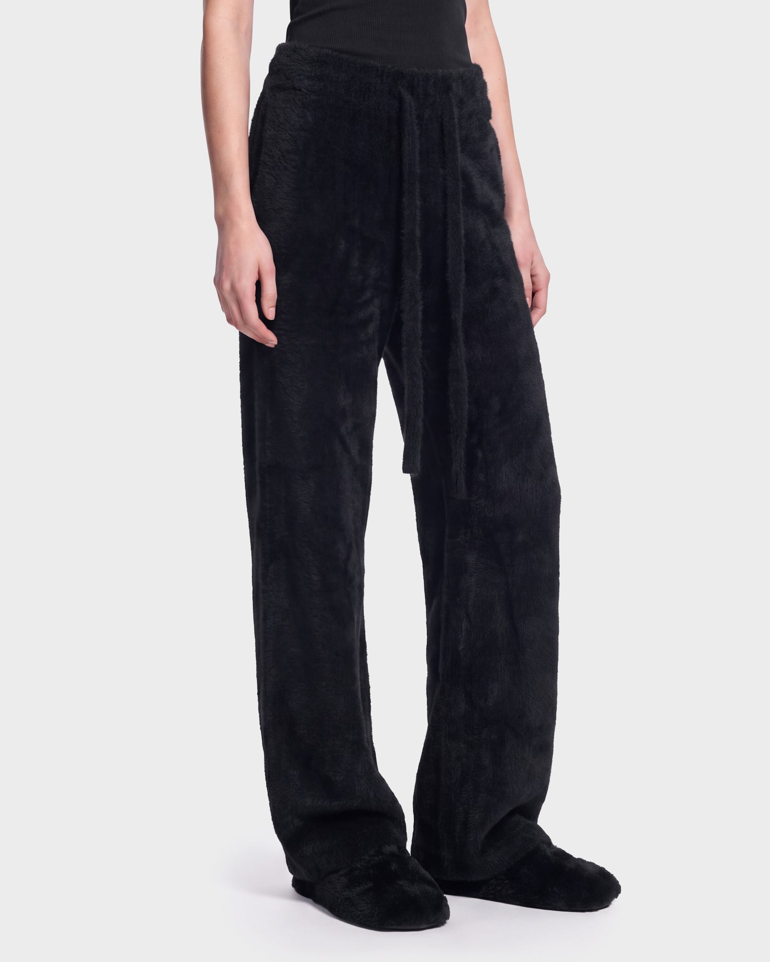 A person is shown from the waist down wearing Rudsak’s GENESIS - 8526506 Black pants—loose-fitting, fuzzy black trousers with a matching tie belt—paired with black slippers, standing against a plain, light background.