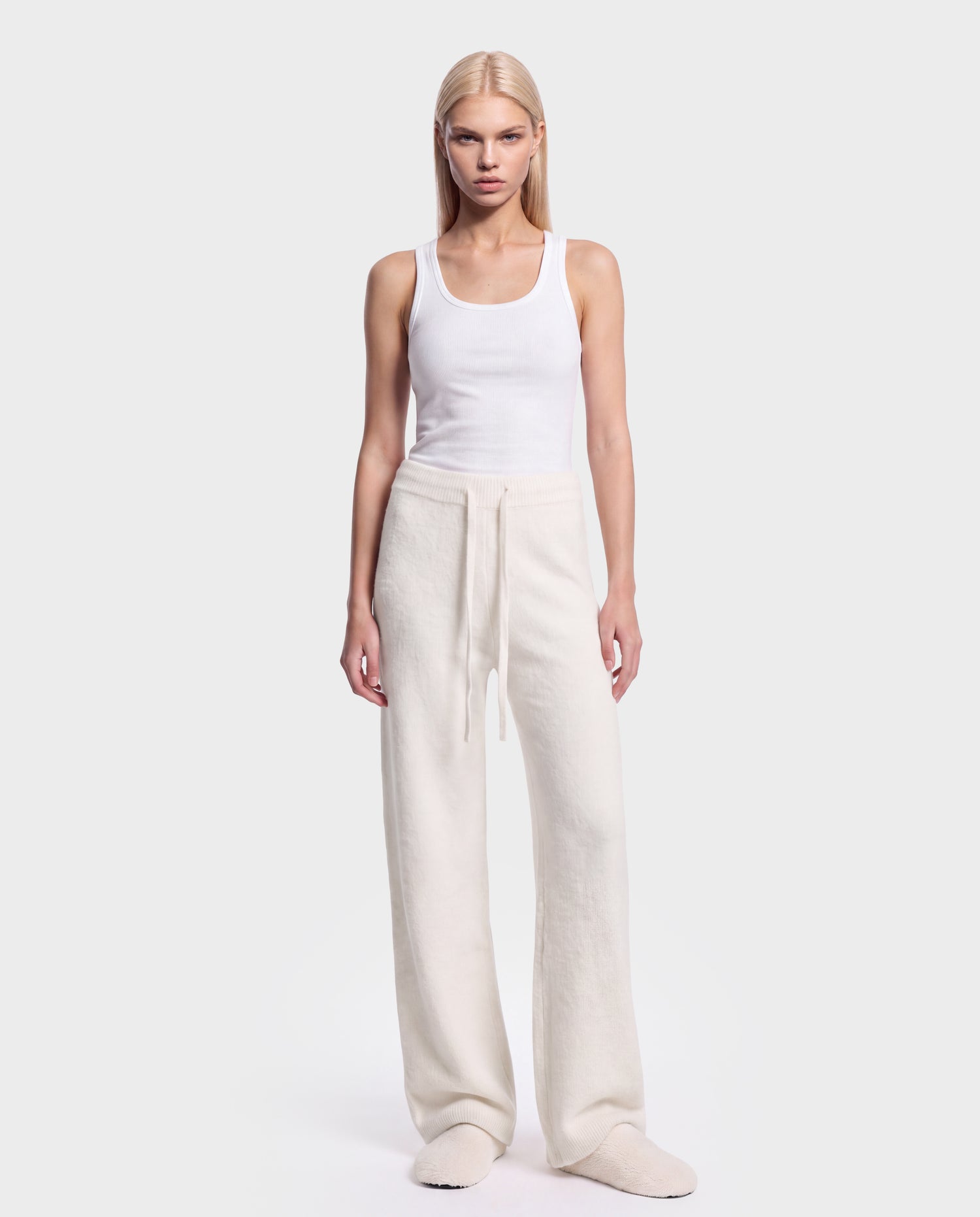 A person stands facing forward against a plain light background, wearing the Rudsak GENESIS - 8526506 Iceland cream-colored loose drawstring pants, paired with a white tank top and cream slippers.
