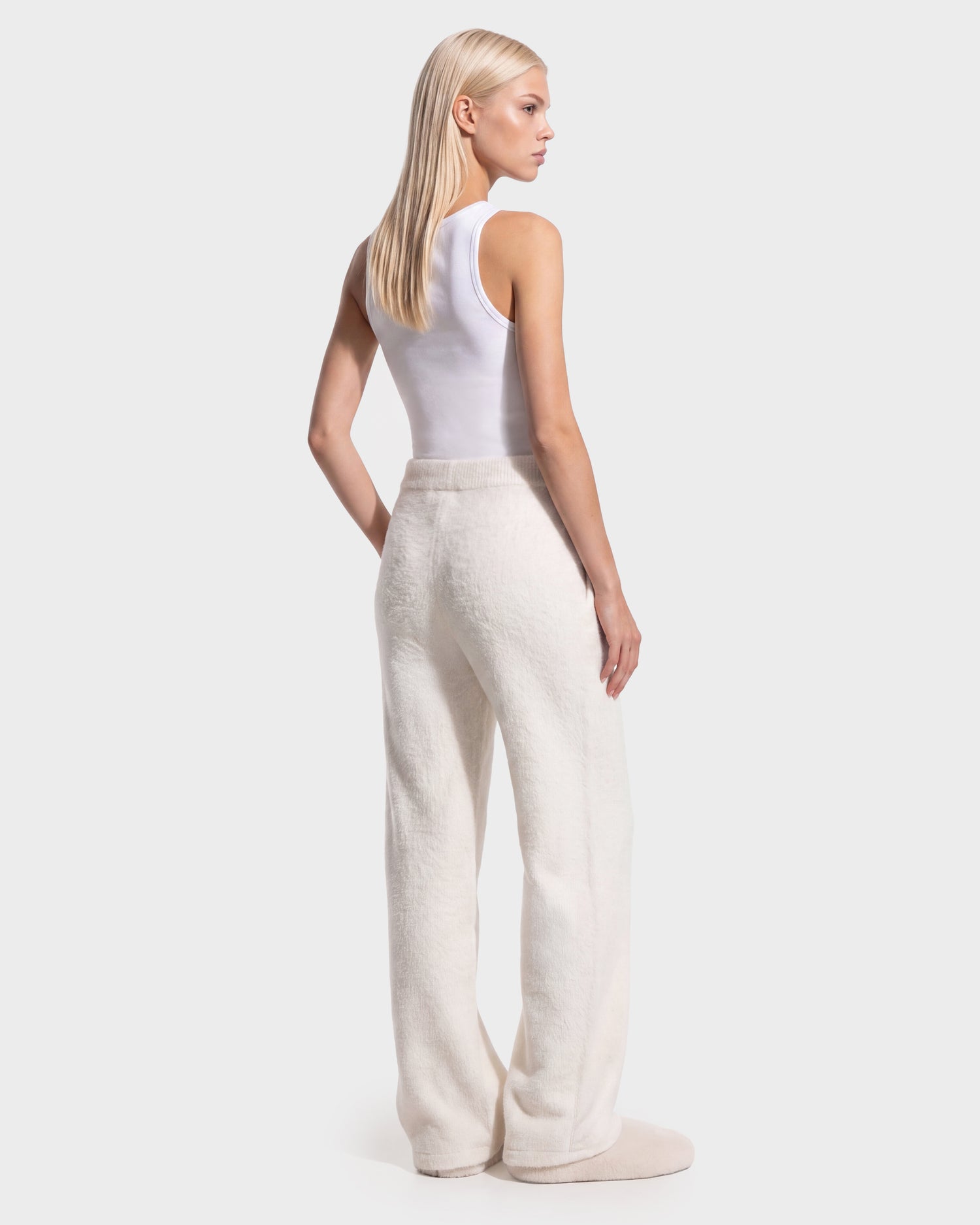 A person with long blonde hair, wearing the Rudsak GENESIS - 8526506 Iceland white sleeveless top and loose white pants, stands facing away with their head turned to the side, posing against a plain light background.