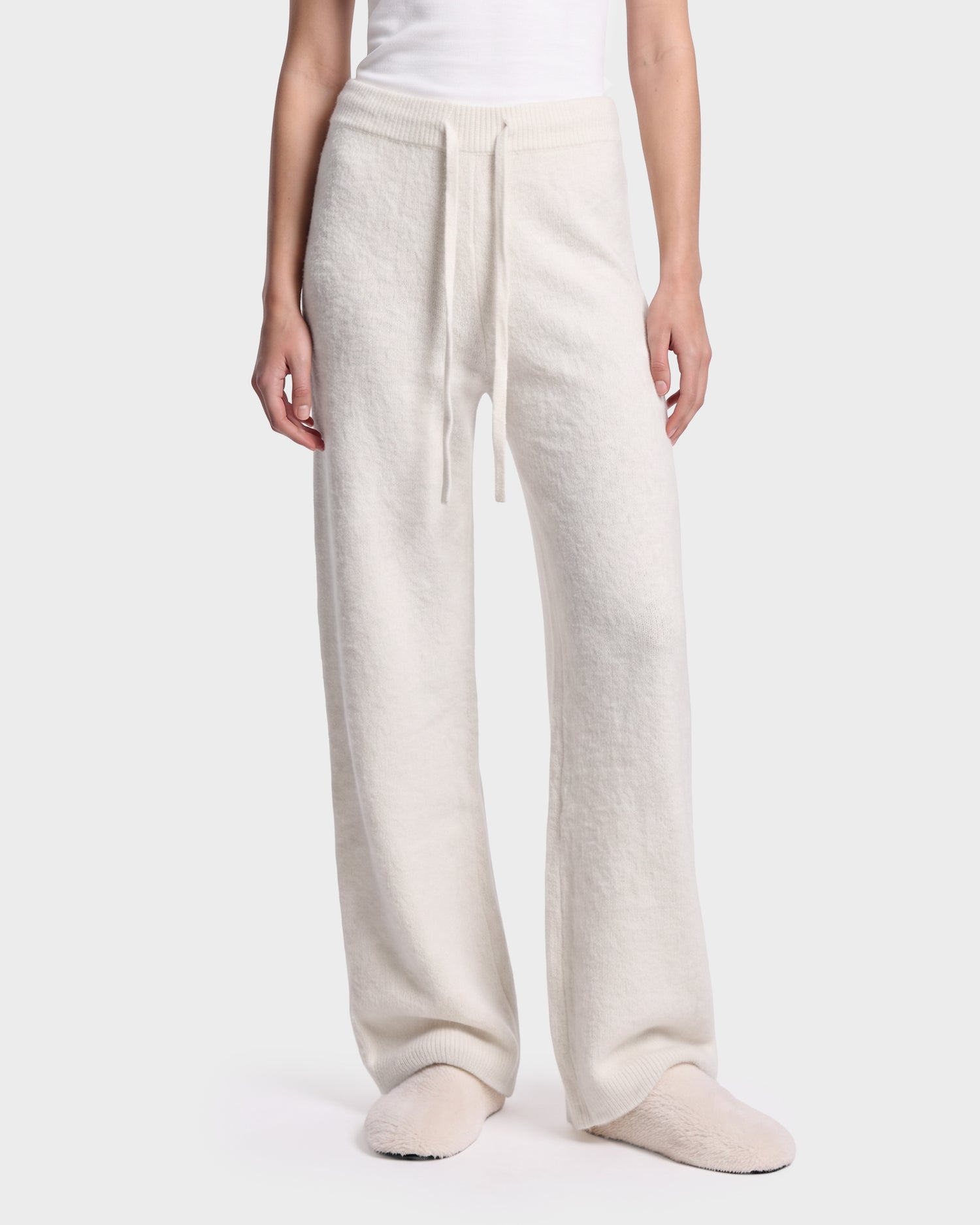 A person wearing the Rudsak GENESIS - 8526506 Iceland loose, cream-colored knit pants with a drawstring waist, paired with matching slippers and a white top, stands against a plain light background.