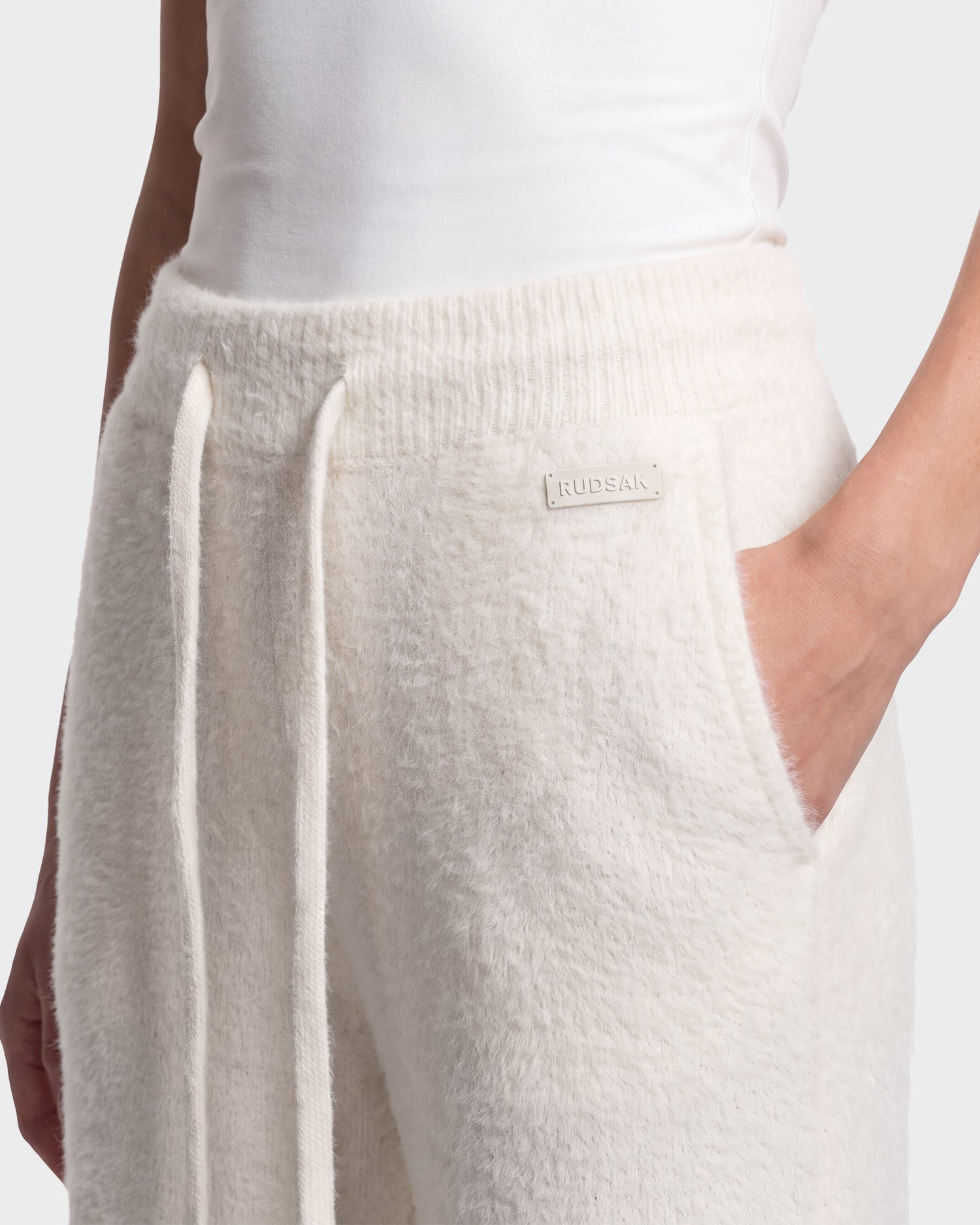 A person wearing soft, white, textured GENESIS - 8526506 Iceland sweatpants by Rudsak with a drawstring waist and hand in pocket, paired with a white tank top. The sweatpants feature a small patch that reads Rudsak.