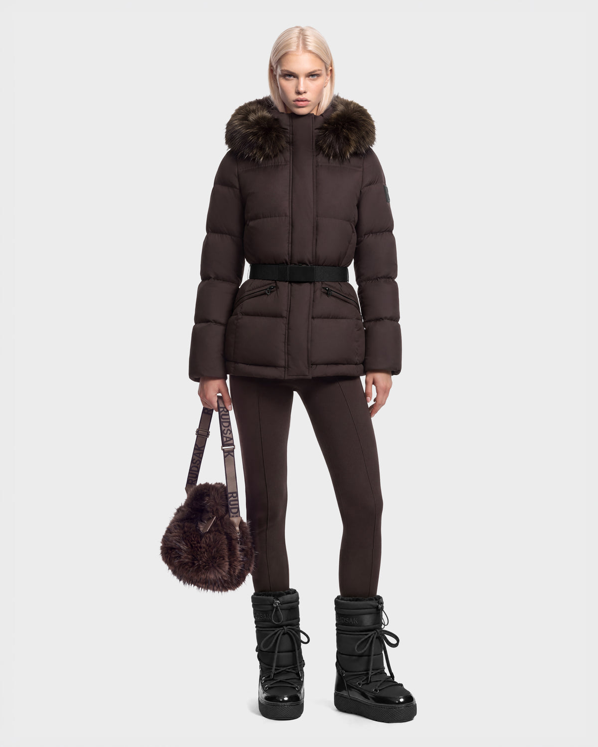 Model wearing a brown Rudsak GRACE FF - 8126535 Espresso goose down puffer coat with a faux-fur trimmed hood and a belted waist, paired with matching leggings, black winter boots, and a furry handbag.