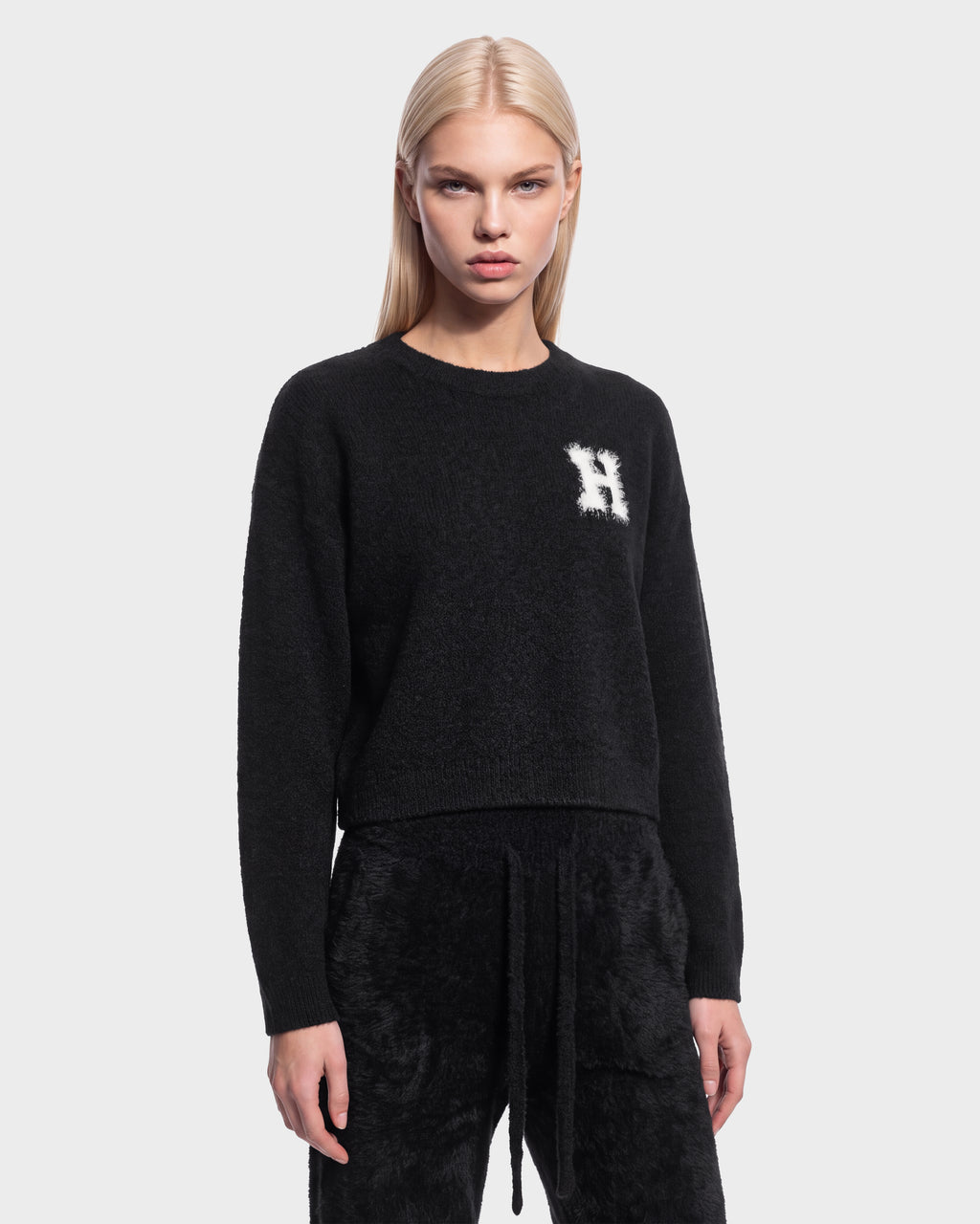 A woman with straight blonde hair wears the black Rudsak HALLIE HV sweater (H526506), featuring a white ‘H’ on the chest, along with matching black pants, as she stands against a plain light background.