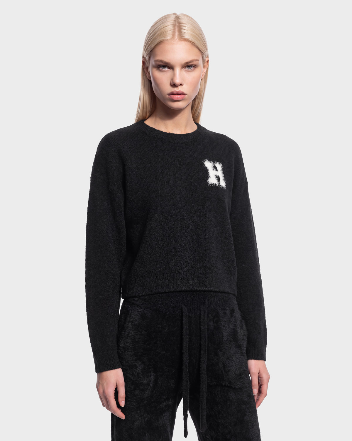 A woman with straight blonde hair wears the black Rudsak HALLIE HV sweater (H526506), featuring a white ‘H’ on the chest, along with matching black pants, as she stands against a plain light background.