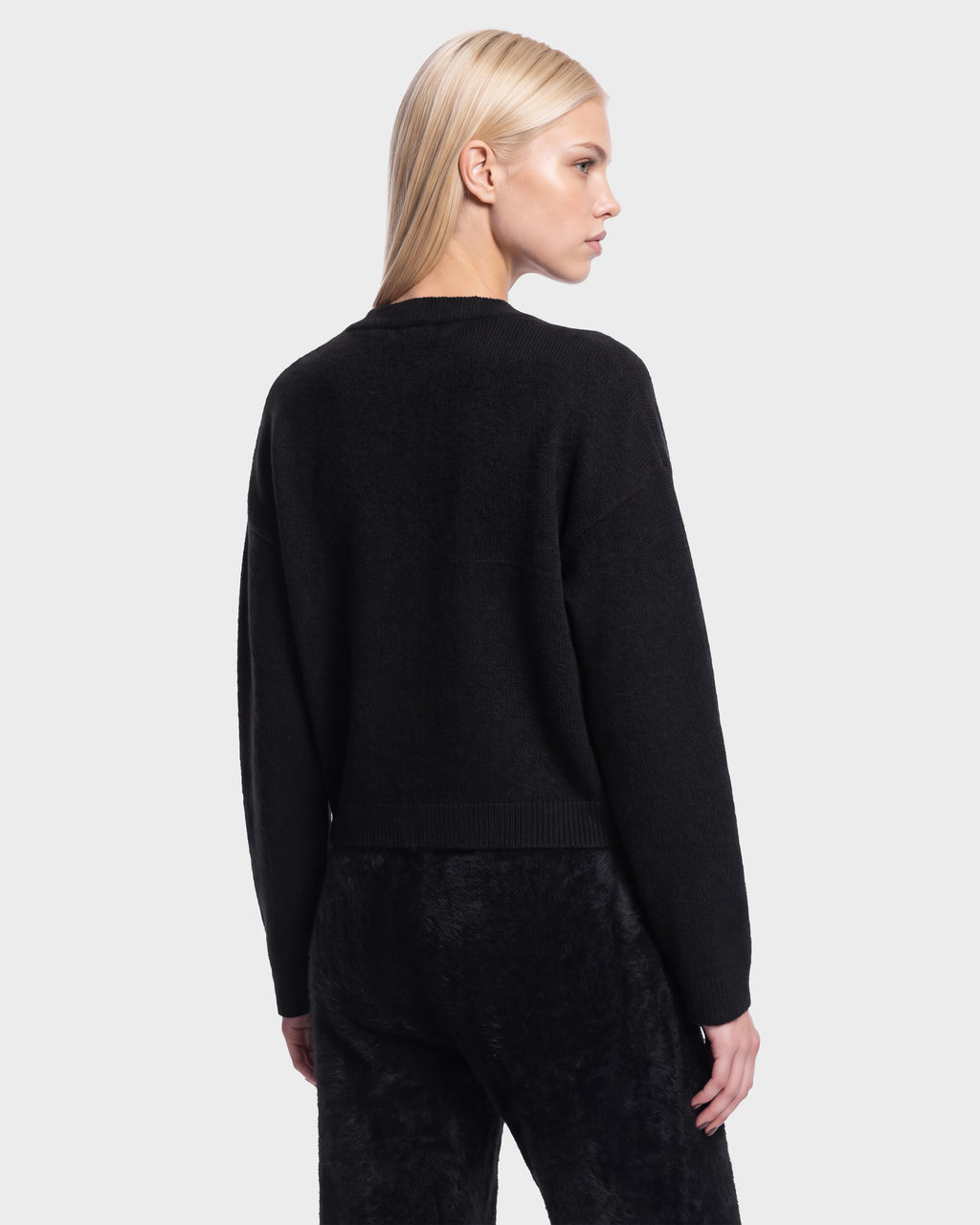 A woman with straight blonde hair stands facing away and glances to the side against a plain light background, wearing the Rudsak HALLIE HV - H526506 crewneck sweater in black paired with black pants.