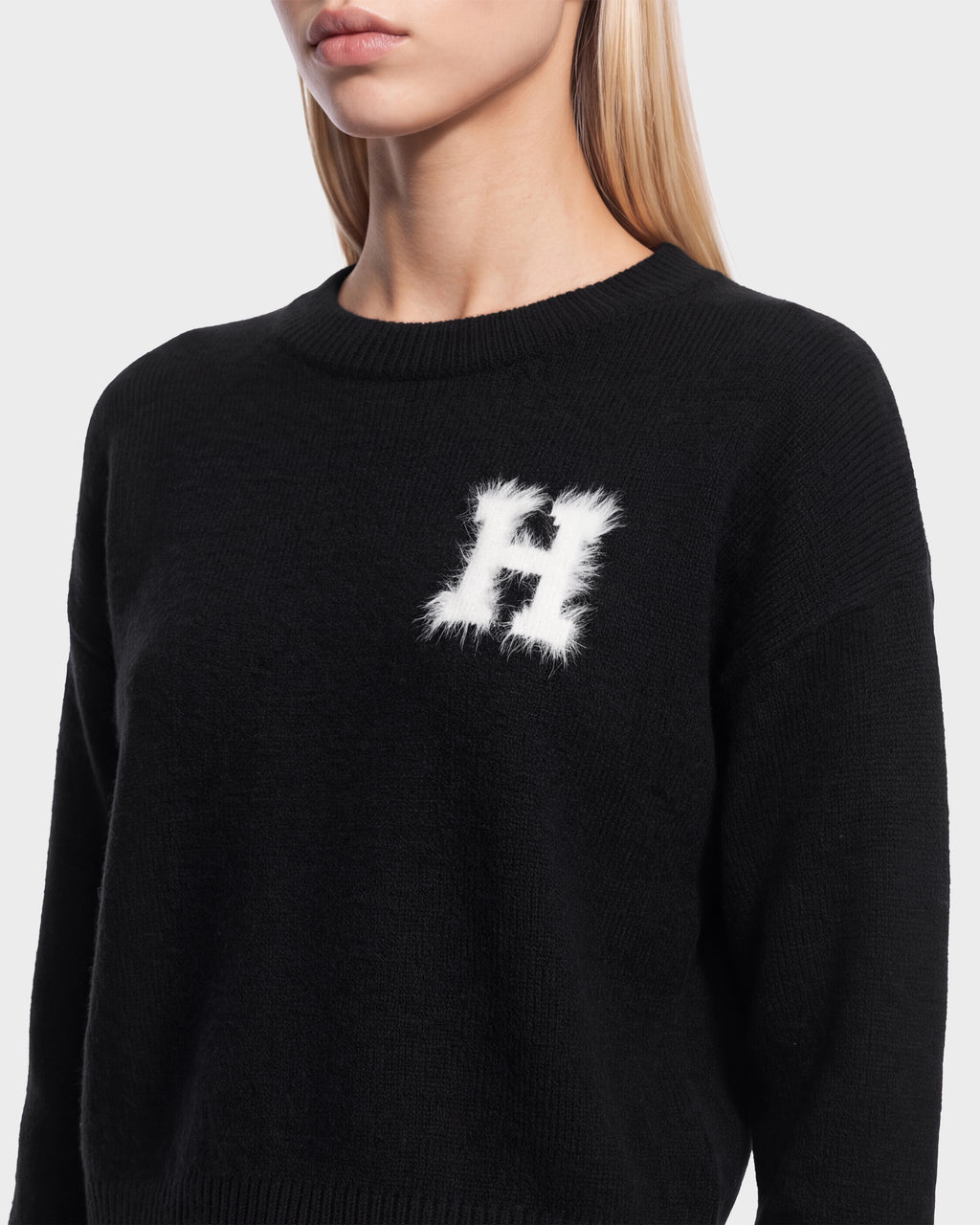 A woman wears the Rudsak HALLIE HV - H526506 Black crewneck sweater featuring a white, fuzzy letter “H” patch on the left chest, evoking classic collegiate style. The background is plain light gray.