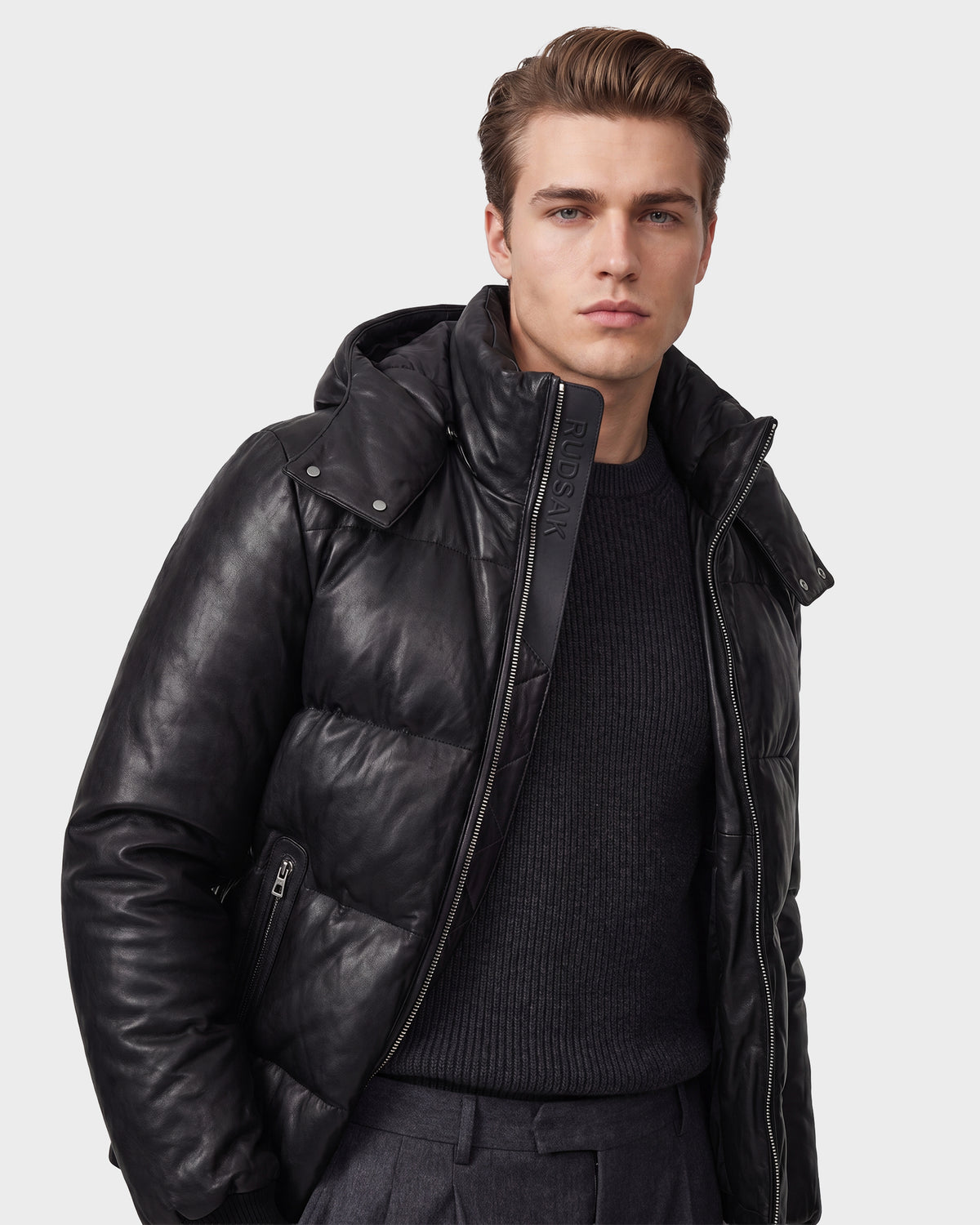 A man with short brown hair is wearing the Rudsak HARVEY X - 6125681 Black puffer jacket over a dark sweater and gray trousers, standing against a plain light background.