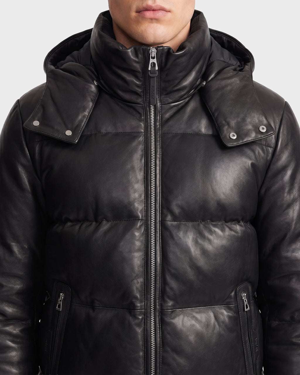 A person wearing the HARVEY X - 6125681 Black, a Rudsak puffer-style quilted leather parka with a high collar and hood, shown from the shoulders to above the mouth, featuring visible zipper and snap details—an iconic piece of Rudsak outerwear.
