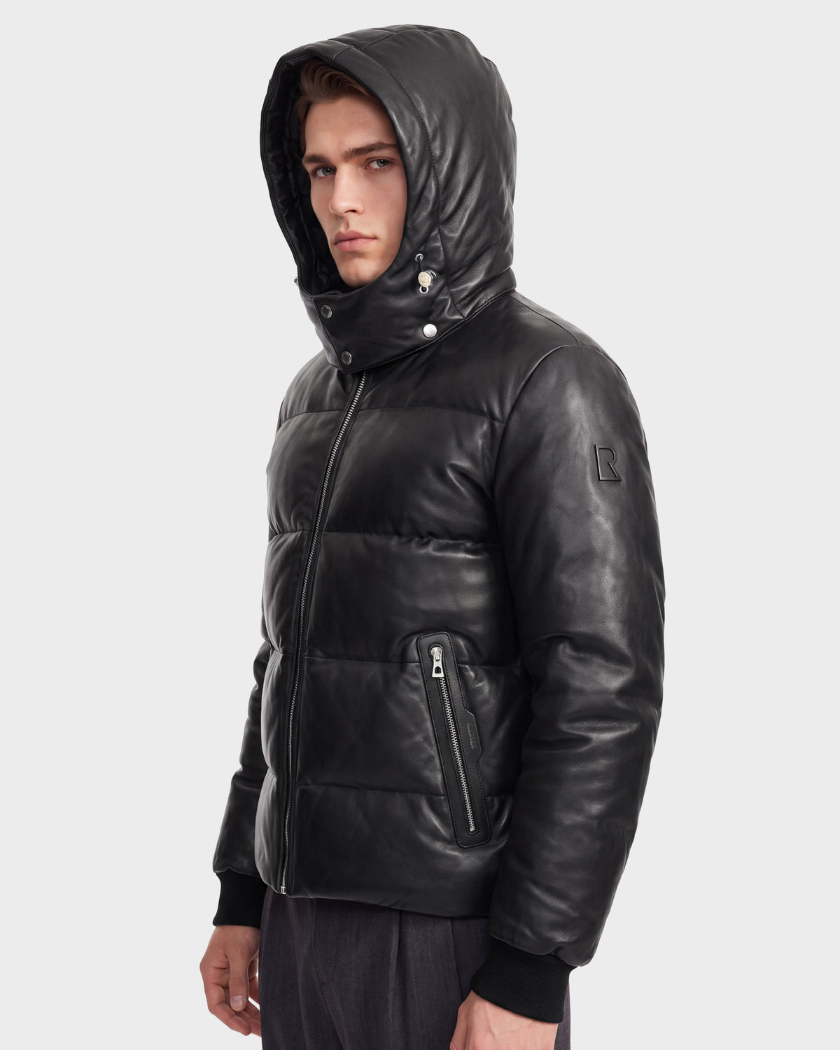 A young man wearing the Rudsak HARVEY X - 6125681 Black hooded quilted leather parka stands facing slightly left, gazing at the camera with a serious expression against a plain light background.