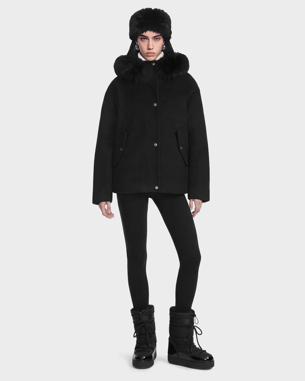 A person wearing a black Rudsak HAZEL - 8126981 wool-blend winter coat with a detachable faux-fur hood, a matching fur hat, leggings, and black insulated winter boots.