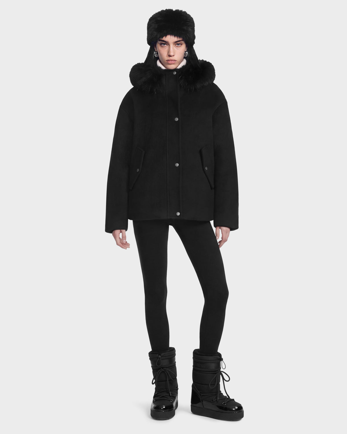 A person wearing a black Rudsak HAZEL - 8126981 wool-blend winter coat with a detachable faux-fur hood, a matching fur hat, leggings, and black insulated winter boots.