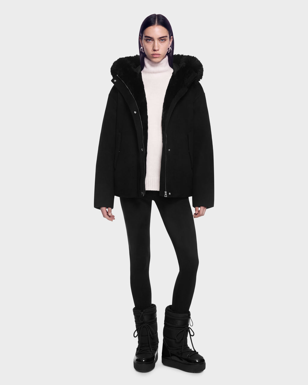 A model wears a black Rudsak HAZEL - 8126981 Black hooded jacket with a detachable faux-fur hood over a white turtleneck sweater, black leggings, and black winter boots.