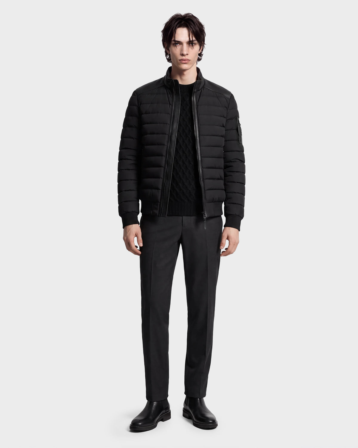 A man stands facing forward against a plain white background, wearing the Rudsak JOSHUA - 6125722 Black quilted down jacket with a leather collar, paired with a black sweater, dark trousers, and black shoes.