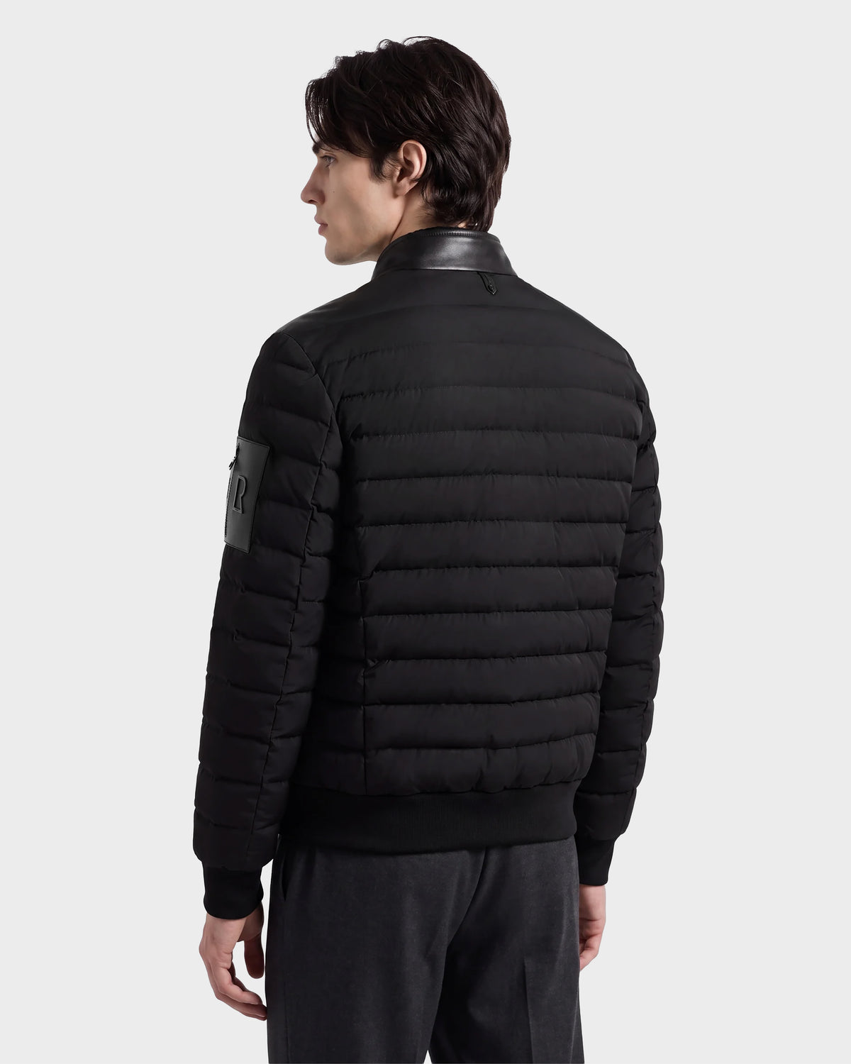 A person with dark hair wears the Rudsak JOSHUA - 6125722 Black quilted down bomber jacket with a leather collar and dark pants, standing and facing away, looking to the left against a plain, light gray background.
