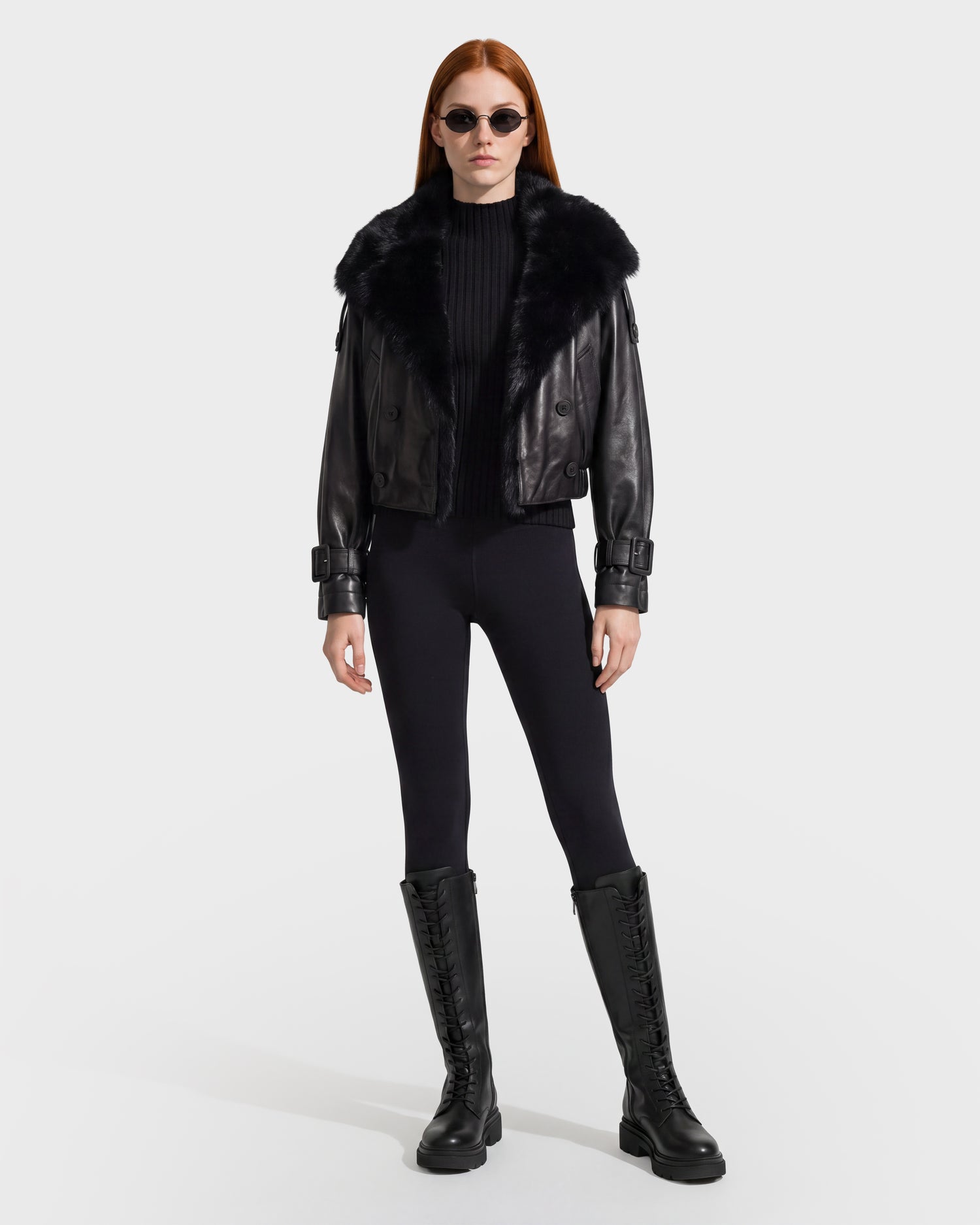 A woman with long red hair wears the Rudsak JANELLE - 8126936 Black fur-collared leather jacket, along with black sunglasses, a black ribbed sweater, black leggings, and tall black lace-up boots, standing against a plain white background.