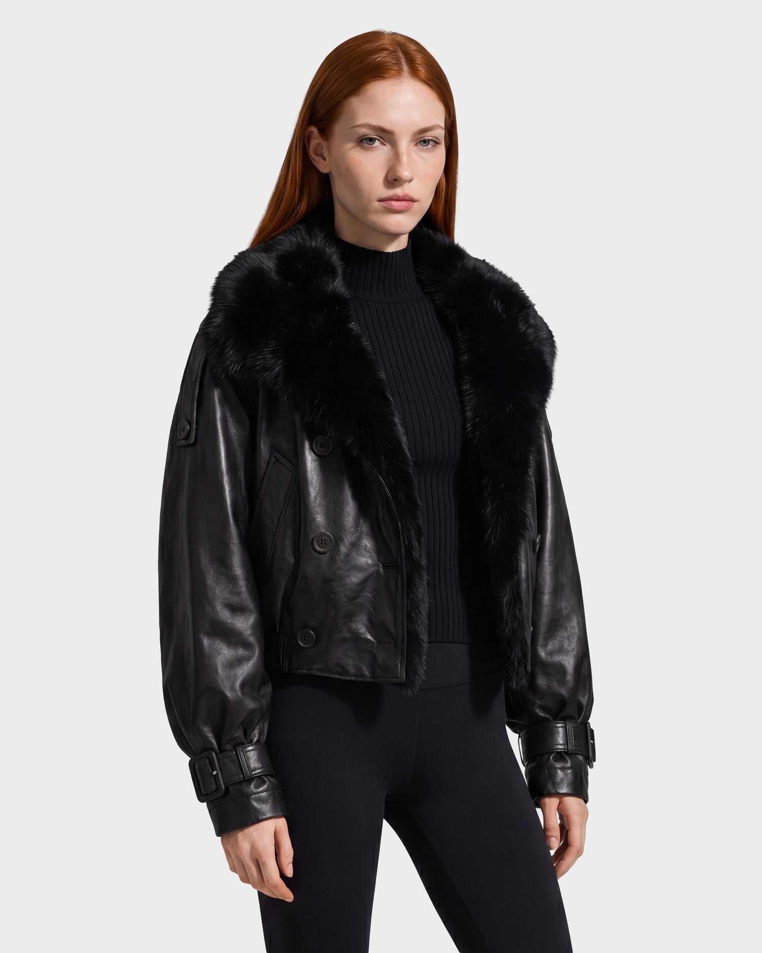 A woman with long red hair wears the Rudsak JANELLE - 8126936 Black leather jacket with a large faux fur collar, styled with a black ribbed sweater and black leggings, standing against a plain light background.