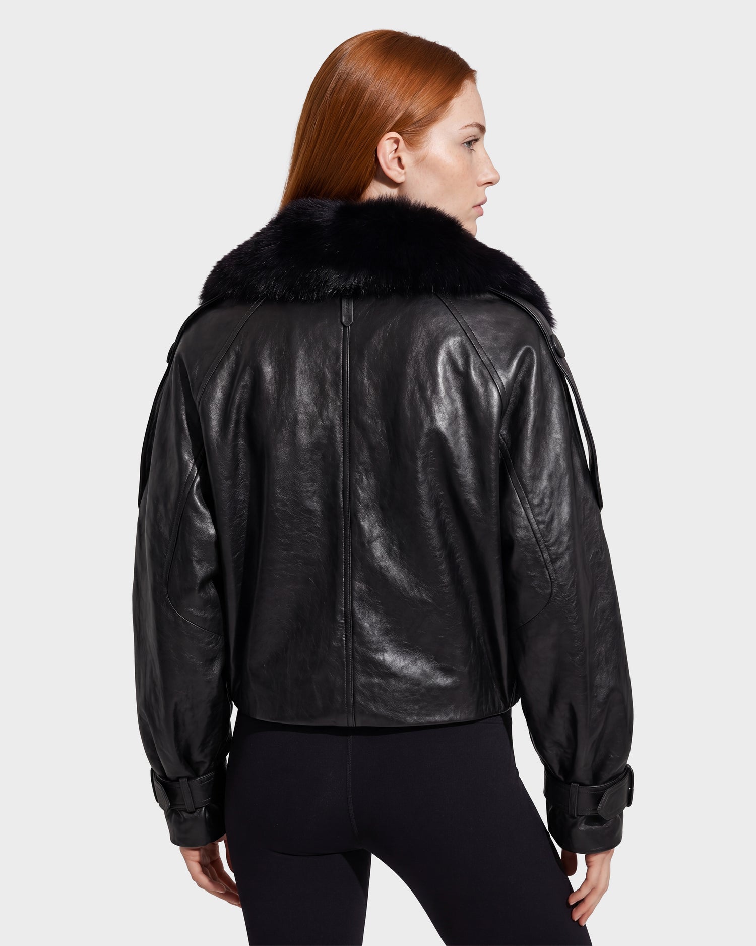 A person with long red hair, wearing the Rudsak JANELLE - 8126936 Black leather jacket with a fur collar, stands facing away from the camera against a plain light background.