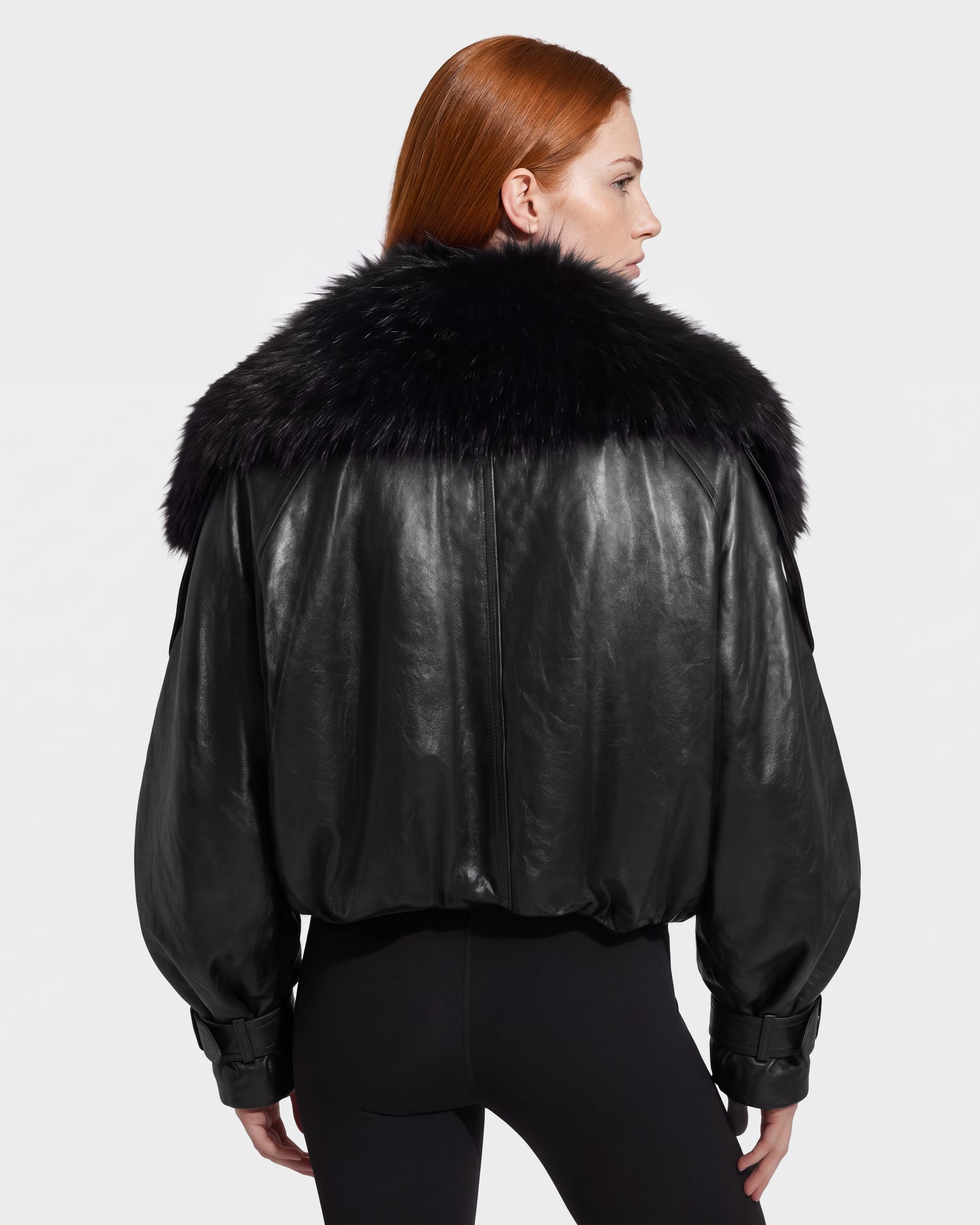 A person with long red hair, seen from the back, is wearing the Rudsak JANELLE - 8126936 Black leather bomber jacket with a large, fluffy faux fur collar and black pants against a plain light background.