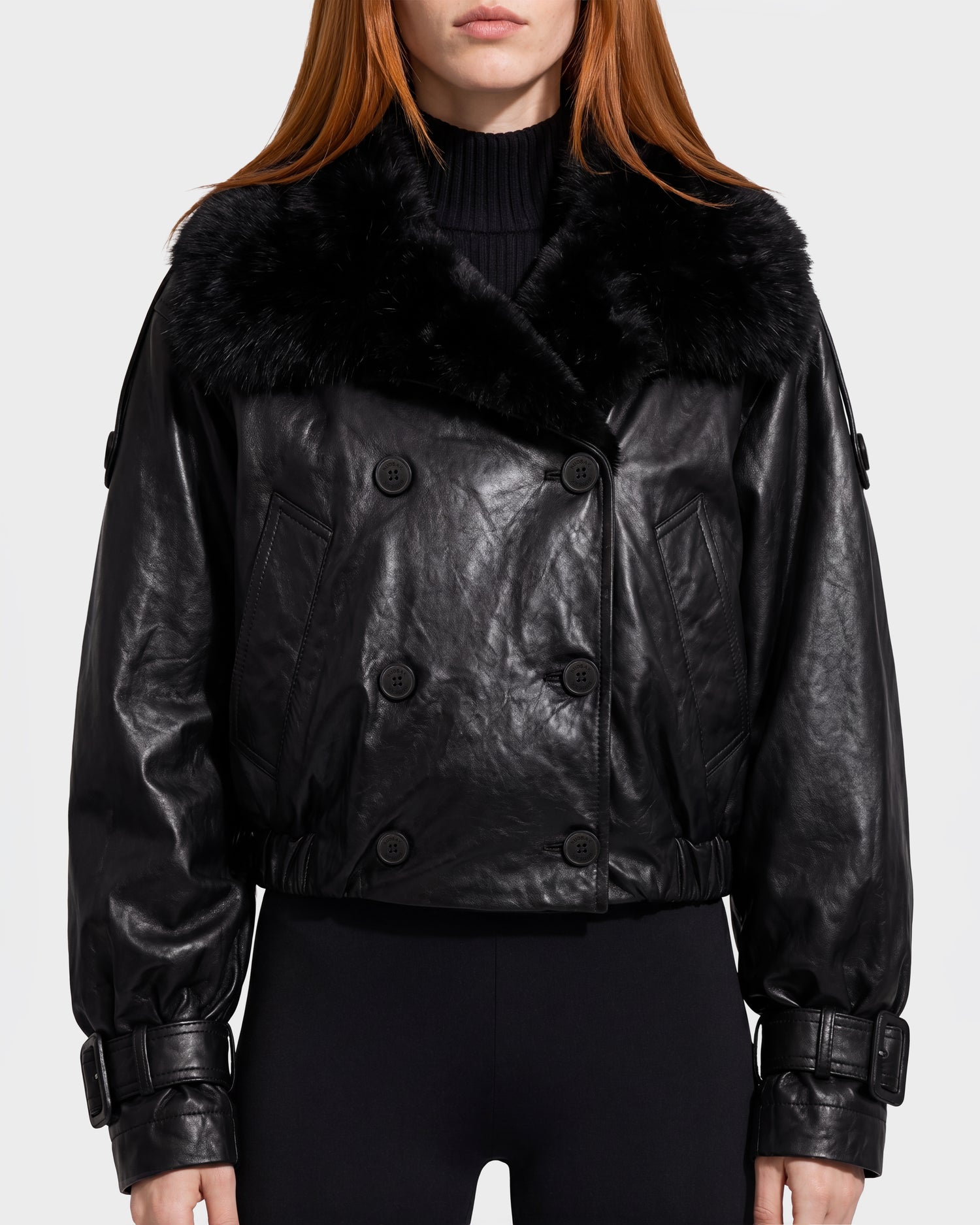 A person wearing the Rudsak JANELLE - 8126936 Black leather jacket, featuring a large black fur collar, double-breasted buttons, and buckled cuffs, paired with black pants. The background is plain and light-colored.