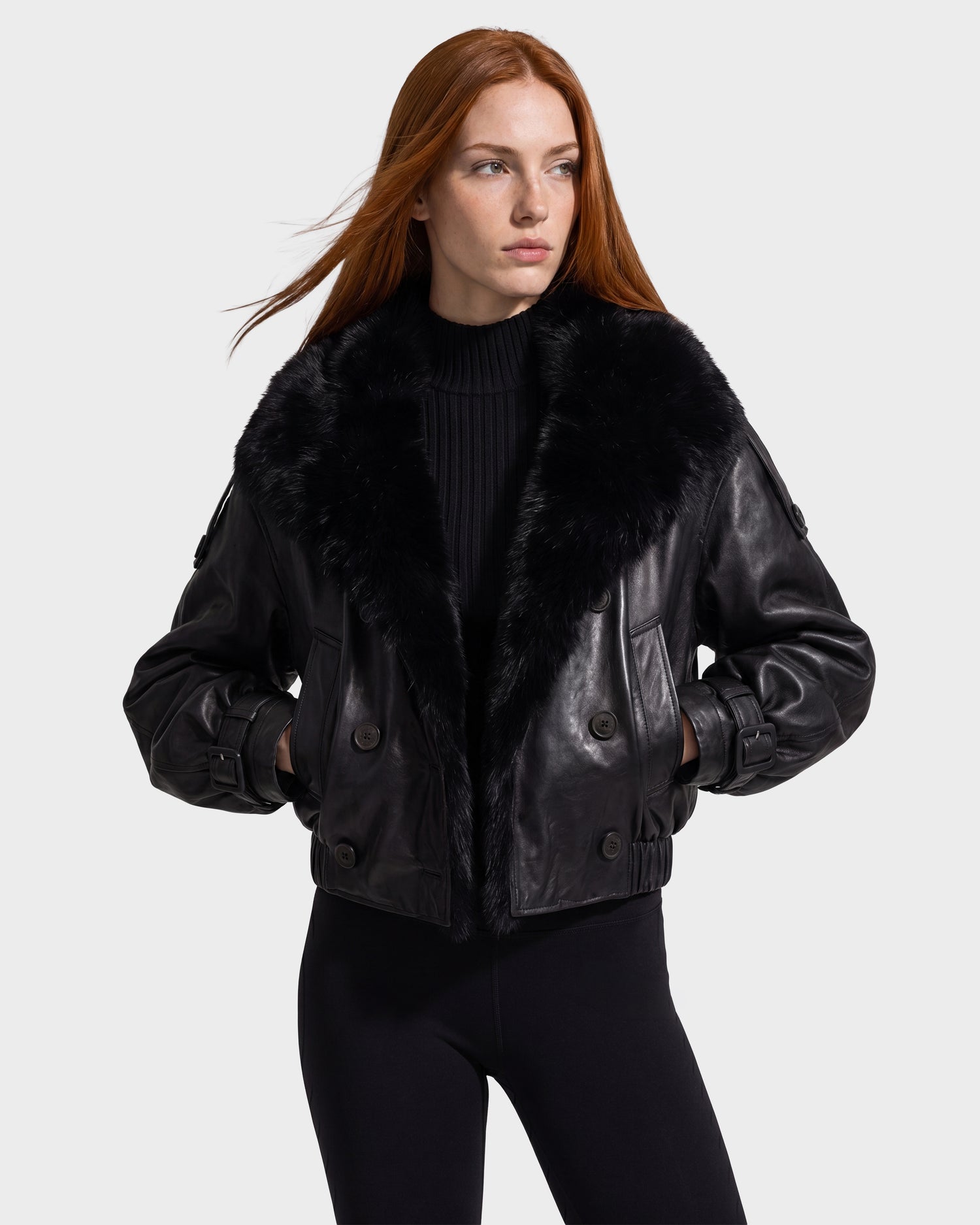 A woman with long red hair is wearing the Rudsak JANELLE - 8126936 Black leather jacket, featuring a large black fur collar, along with a black top and black pants, as she stands with her hands in her pockets against a light gray background.