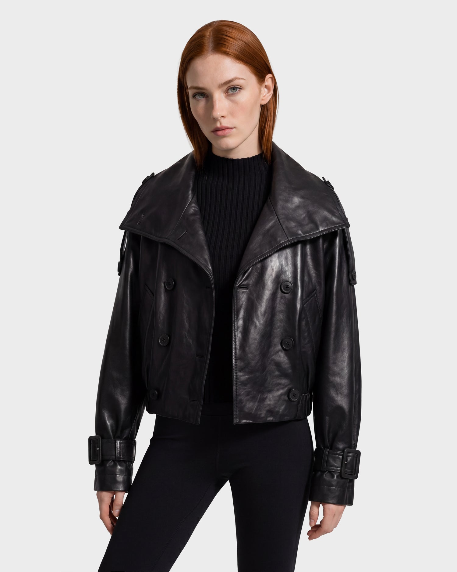 A woman with straight red hair models the Rudsak JANELLE - 8126936 Black leather double-breasted jacket, featuring large lapels and belted cuffs, layered over a black ribbed sweater against a plain light gray background.