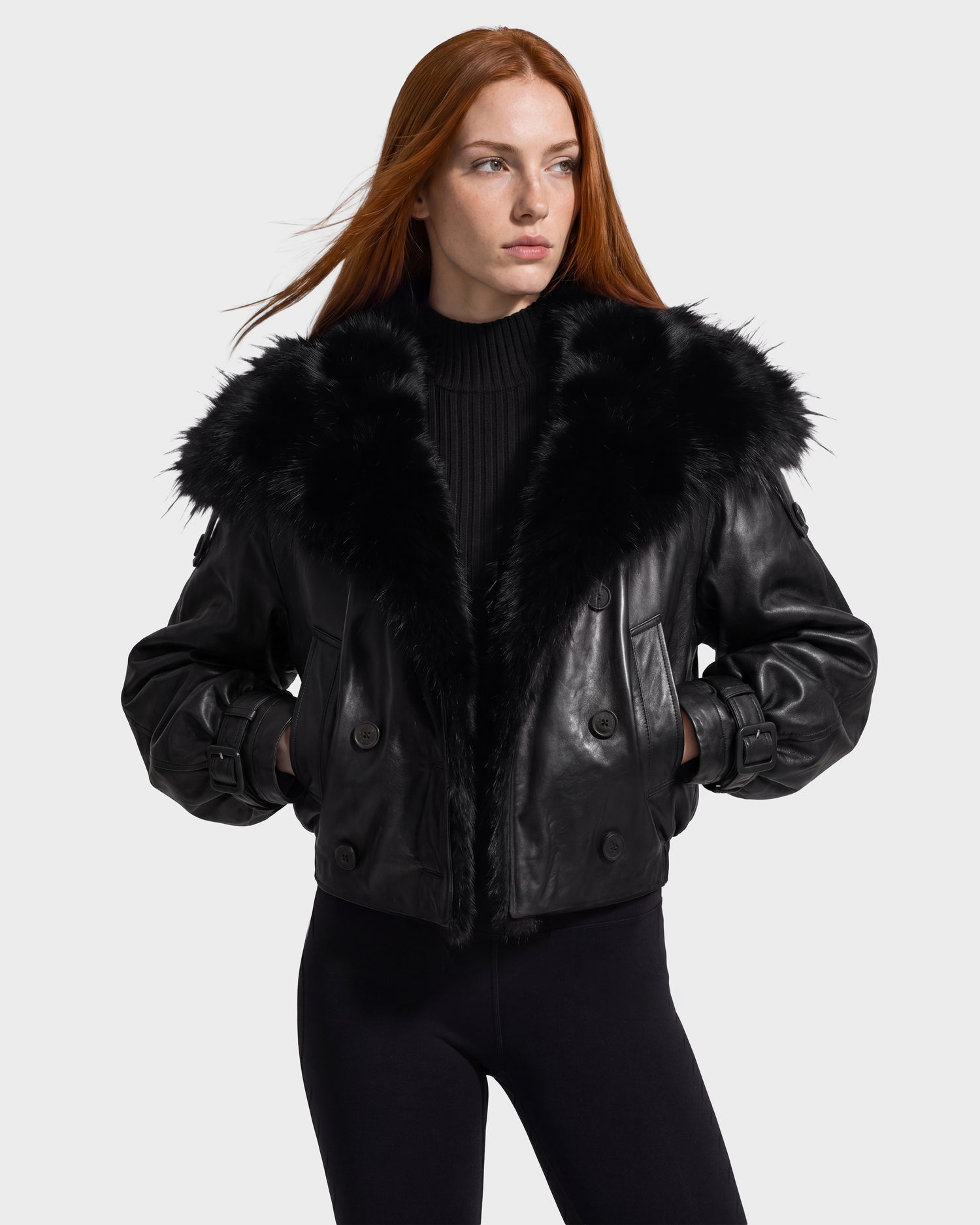 A woman with long red hair wears the Rudsak JANELLE - 8126936 Black leather bomber jacket featuring a large faux fur collar, paired with a black sweater and black pants, standing against a plain light background.