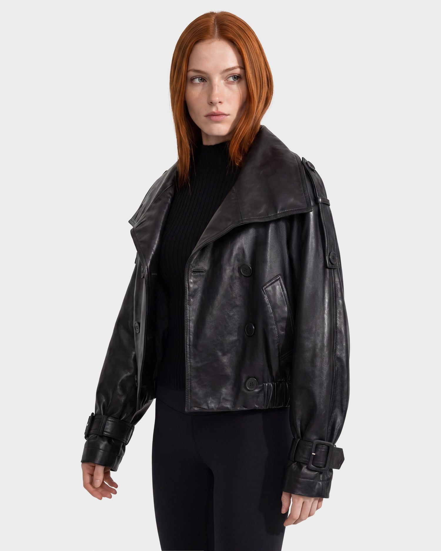A woman with straight red hair wears the Rudsak JANELLE - 8126936 black leather jacket and black pants, standing against a plain light gray background and looking slightly to her left.