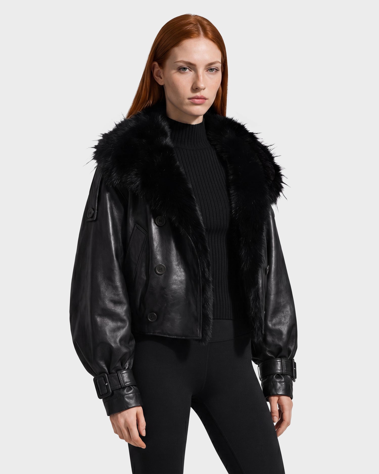 A woman with long red hair wears the Rudsak JANELLE - 8126936 Black jacket—a stylish black leather bomber featuring a large faux fur collar—paired with a black ribbed turtleneck and black leggings, standing against a plain light background.