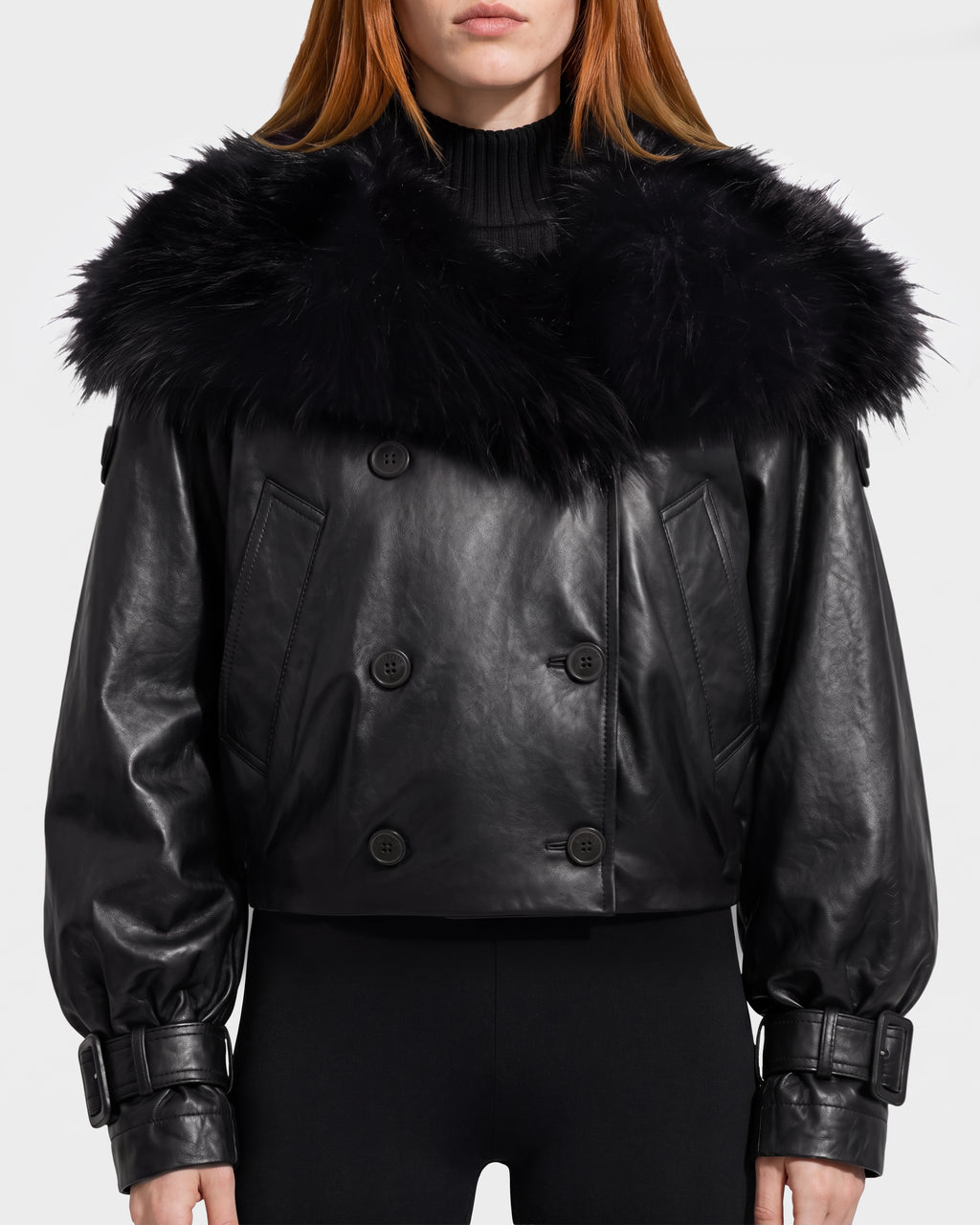 A woman faces forward, showcasing the Rudsak JANELLE - 8126936 Black jacket—a stylish women’s black leather bomber featuring a large faux fur collar, wide sleeves, buckled cuffs, and black buttons. She pairs it with black pants, her straight reddish-brown hair visible for a chic and modern look.