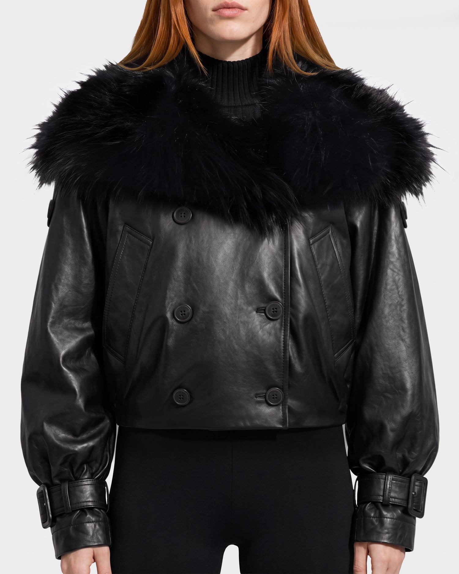 A woman faces forward, showcasing the Rudsak JANELLE - 8126936 Black jacket—a stylish women’s black leather bomber featuring a large faux fur collar, wide sleeves, buckled cuffs, and black buttons. She pairs it with black pants, her straight reddish-brown hair visible for a chic and modern look.