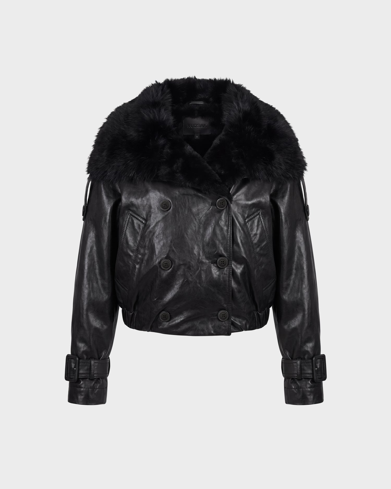 JANELLE - 8126936 Black by Rudsak is a black leather jacket featuring a wide faux fur collar, double-breasted button front, belted cuffs, and a cropped length, displayed on a plain light gray background.