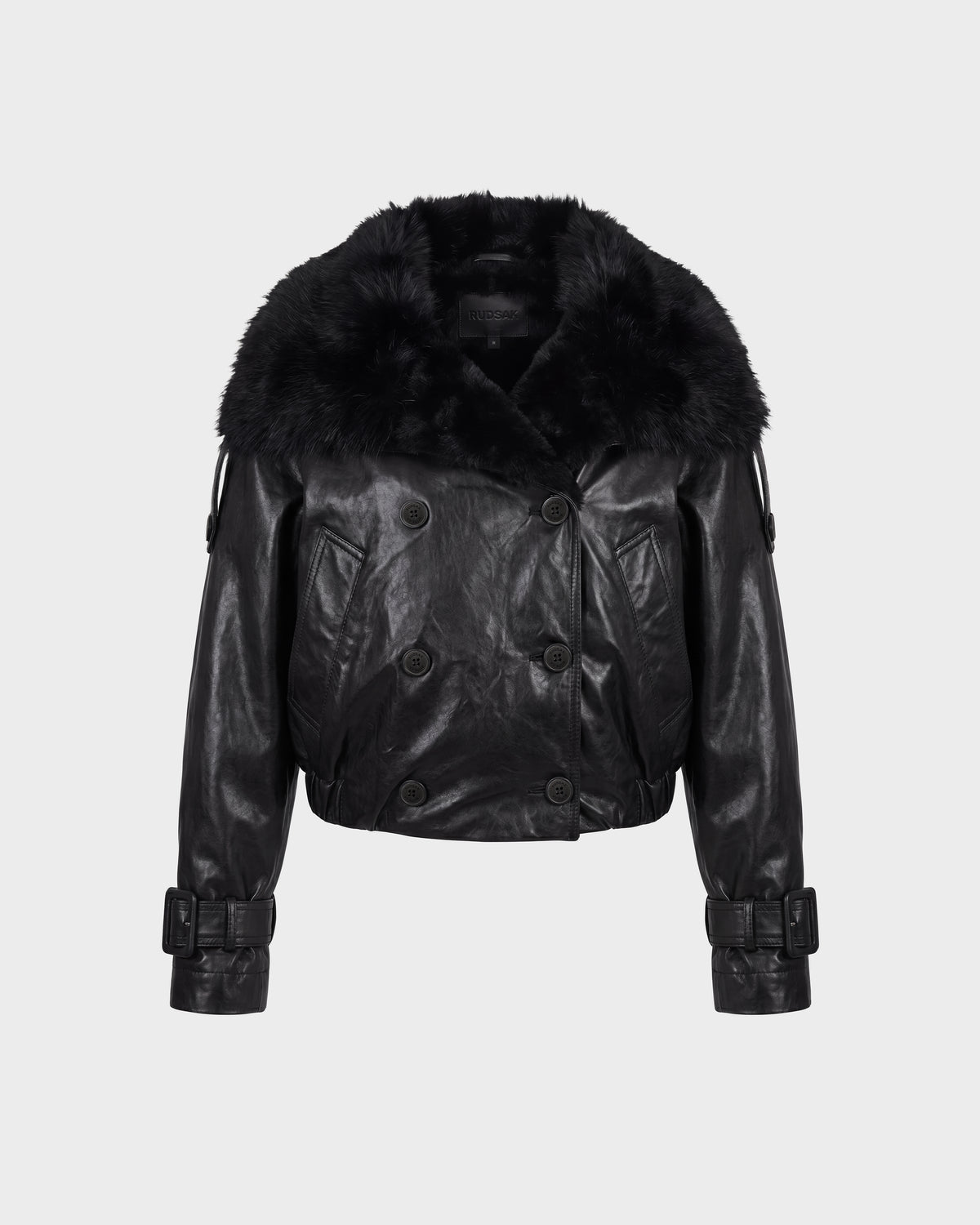 JANELLE - 8126936 Black by Rudsak is a black leather jacket featuring a wide faux fur collar, double-breasted button front, belted cuffs, and a cropped length, displayed on a plain light gray background.