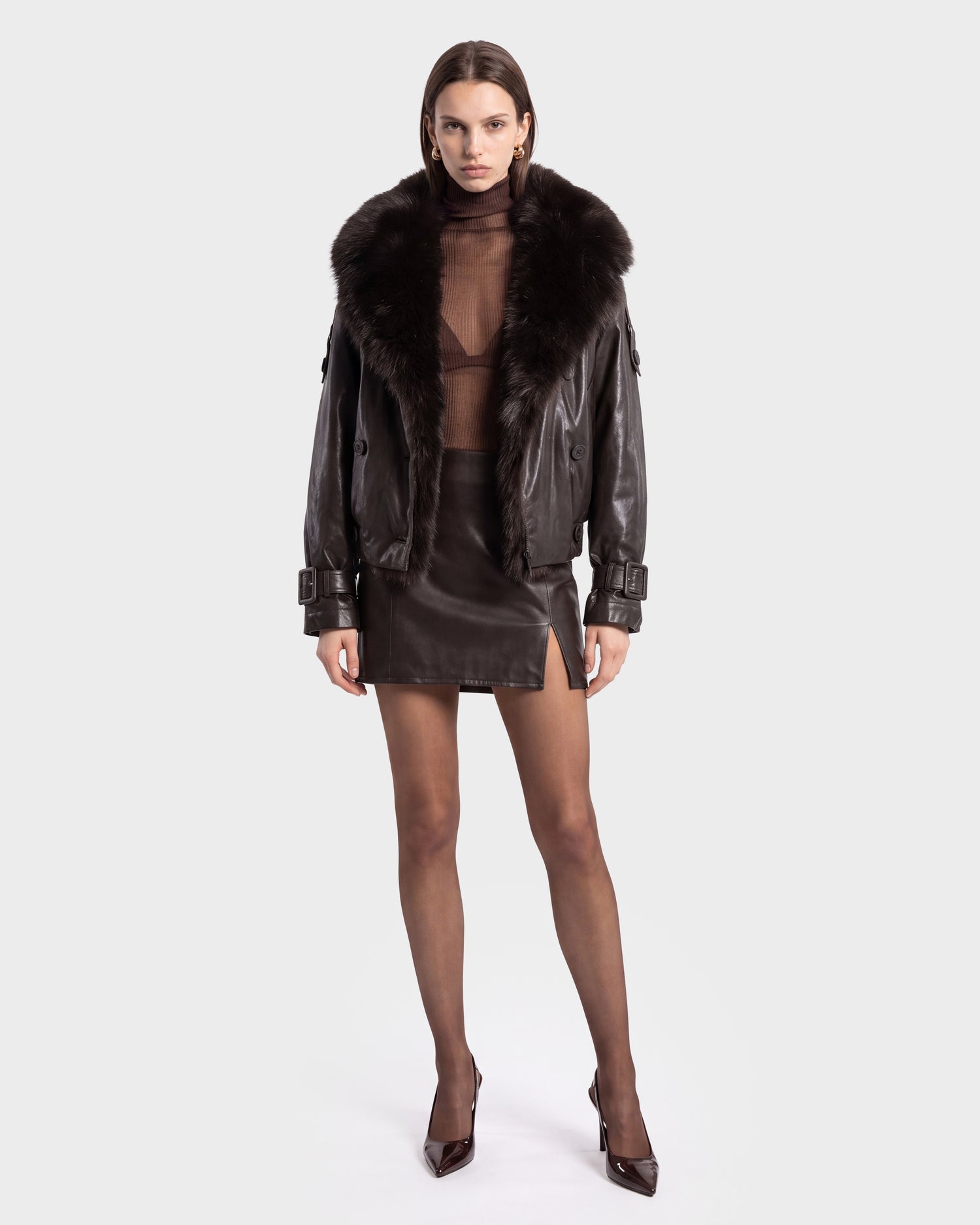 A woman stands facing forward against a plain light background, wearing the Rudsak JANELLE - 8126936 Espresso bomber jacket—a dark leather piece with a faux fur collar—styled over a sheer brown top and black leather miniskirt. She completes her modern silhouette with sheer tights and pointed heels.
